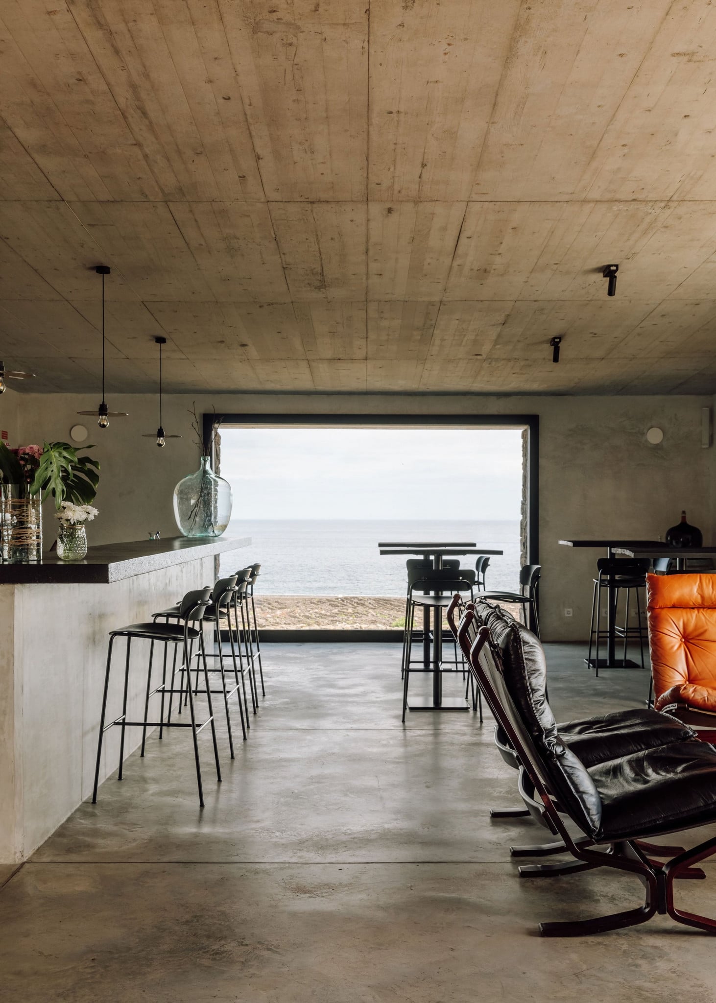 Azores Wine Company, Europe — Concrete coastal bar with ocean views, minimalist black seating, pendant lighting, and floor-to-ceiling windows opening onto beach.