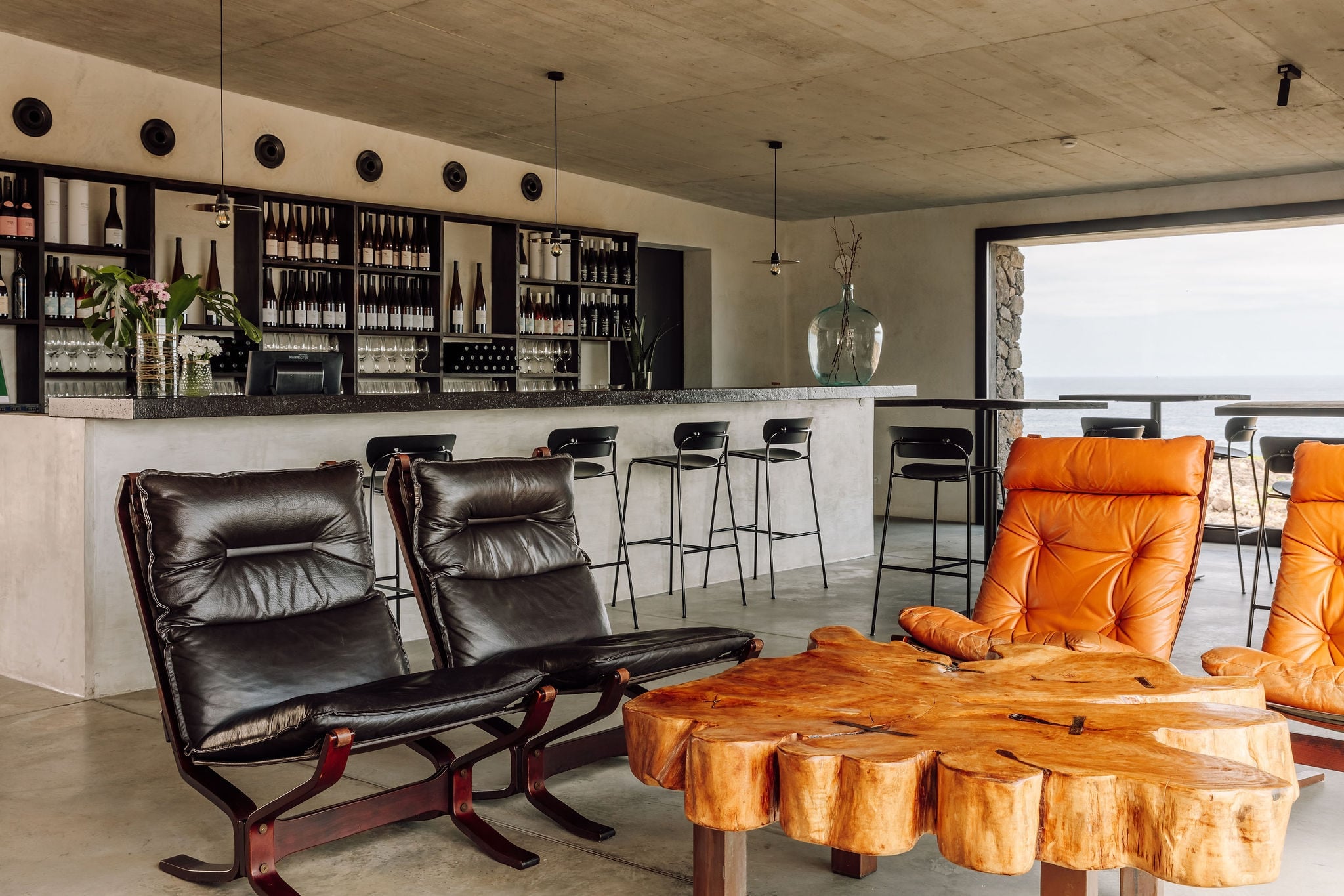 Azores Wine Company, Europe — Modern wine bar lounge with black leather chairs, natural wood slab table, orange cushioned seating, and extensive wine shelving against concrete backdrop.