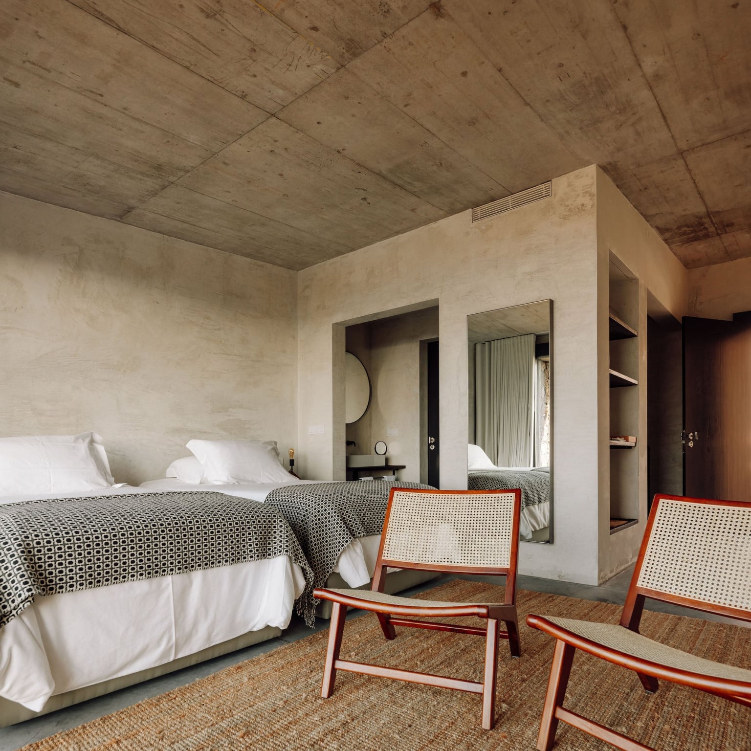 Azores Wine Company, Europe — Minimalist hotel bedroom with concrete ceiling, cream plaster walls, geometric patterned bedding, rattan chairs, and integrated bathroom pod.