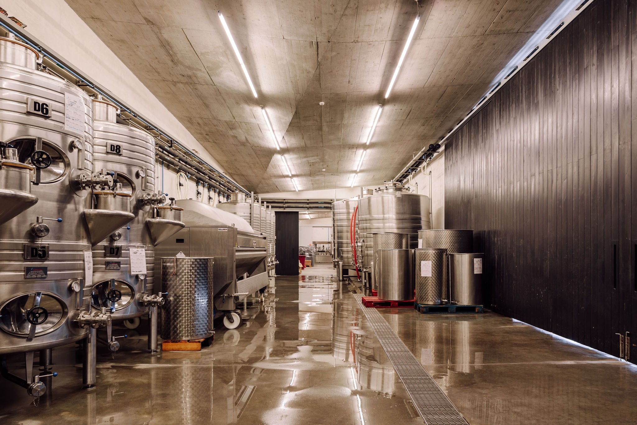 Modern winemaking facility with stainless steel fermentation tanks, polished concrete floors, industrial lighting, and minimalist design aesthetic.