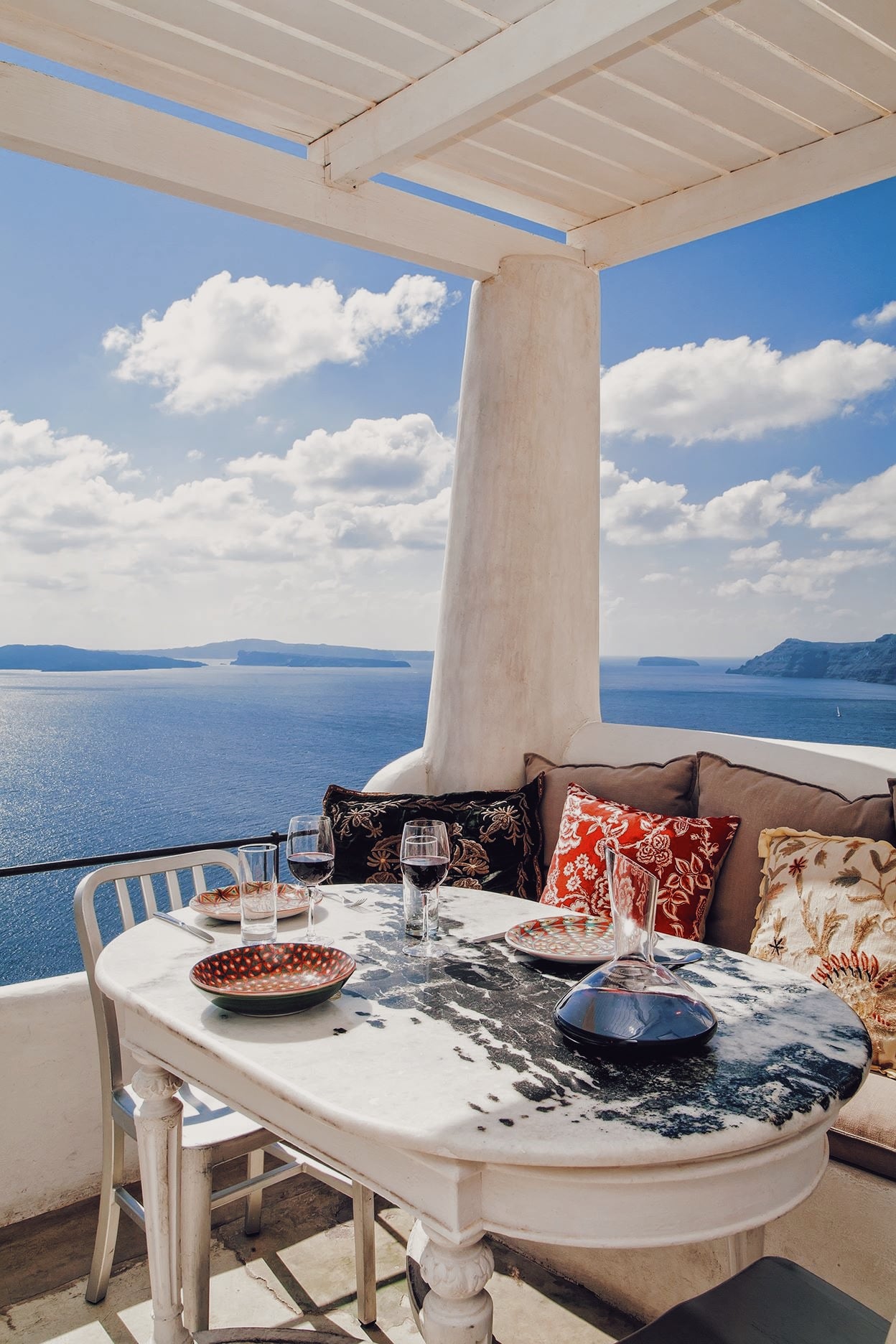 Red Bicycle, Europe — Whitewashed terrace with marble table, colorful pillows, and aegean sea view beneath pergola with supporting column.