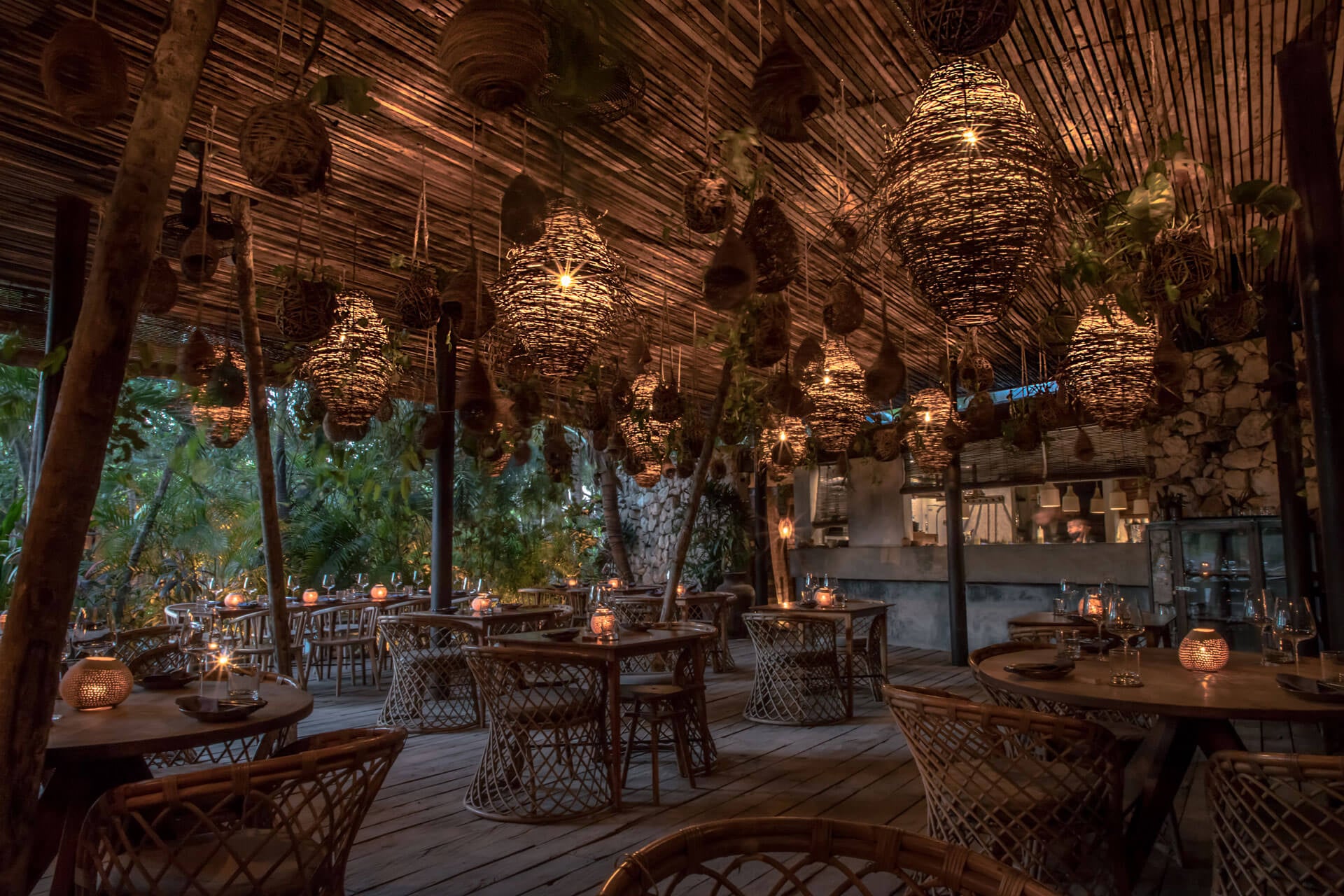 Be Tulum, Mexico — Woven pendant lights illuminate wooden terrace dining pavilion with rattan chairs, tropical trees, and ambient candlelight at dusk.