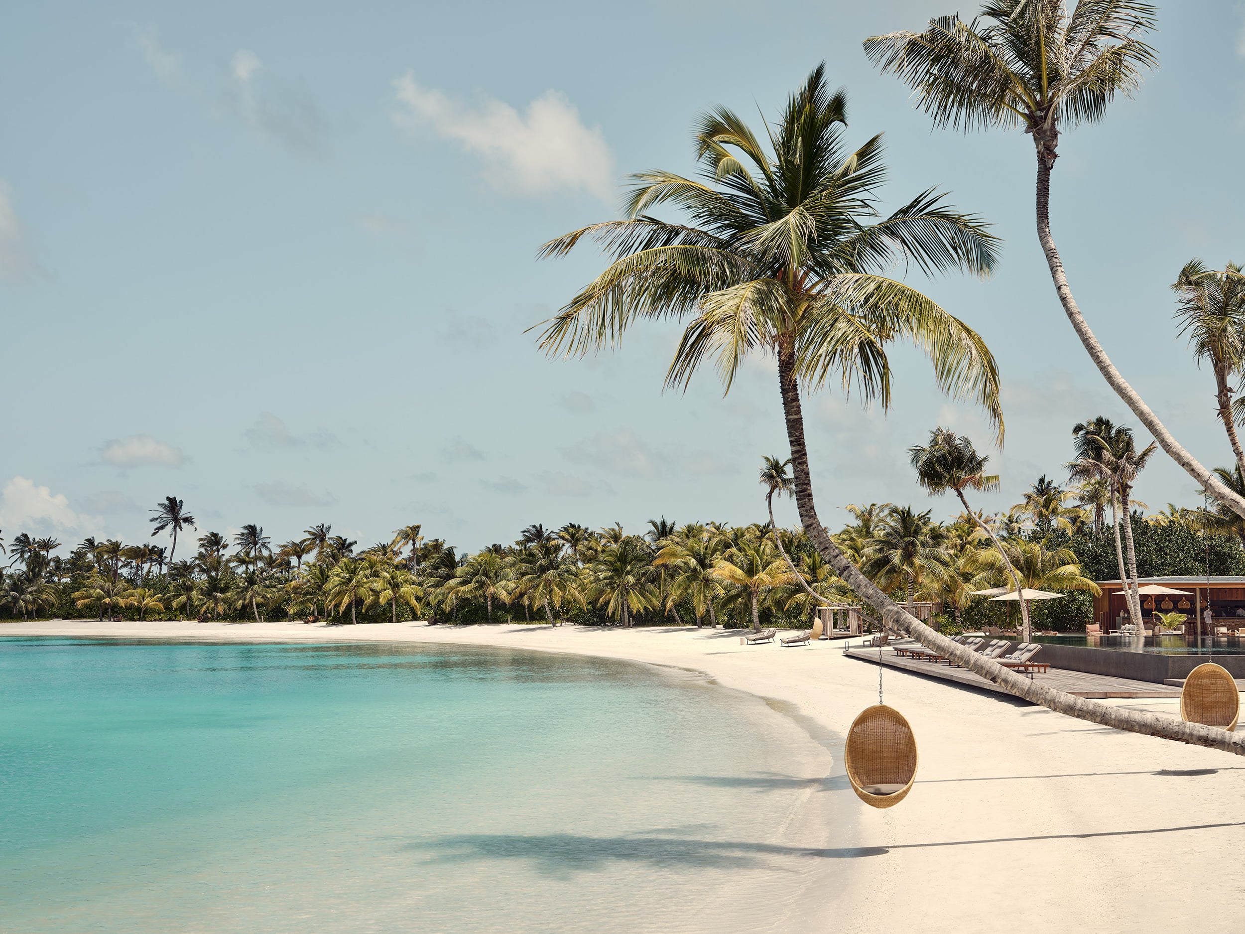 Patina Maldives, Maldives — Turquoise lagoon, white sand beach with suspended woven chairs, curved palm trunk, tropical palms, and thatched beach pavilions under clear sky.
