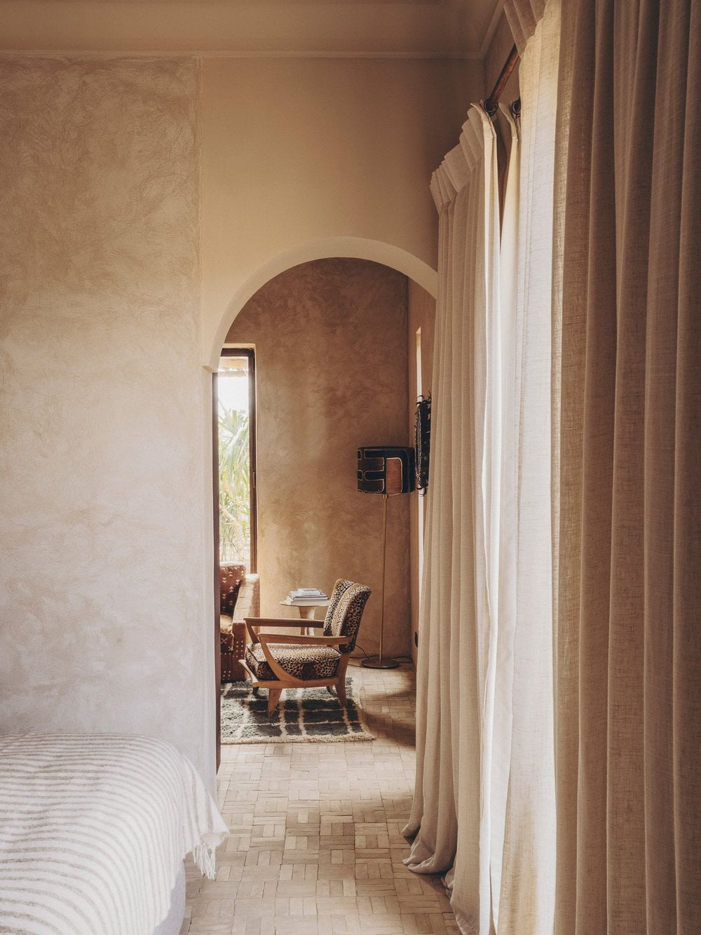 Farasha Farmhouse, Africa — Arched doorway with linen curtains opening to bedroom with leopard-print chair, ochre plaster walls, and natural light through narrow window.