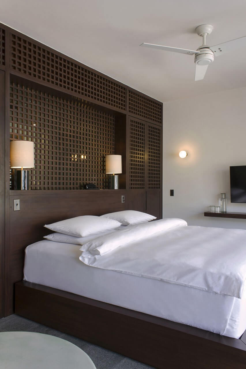 Baja Club, Mexico — Modern hotel bedroom with dark wood lattice headboard, white linens, paired table lamps, ceiling fan, and minimalist coastal design aesthetic.