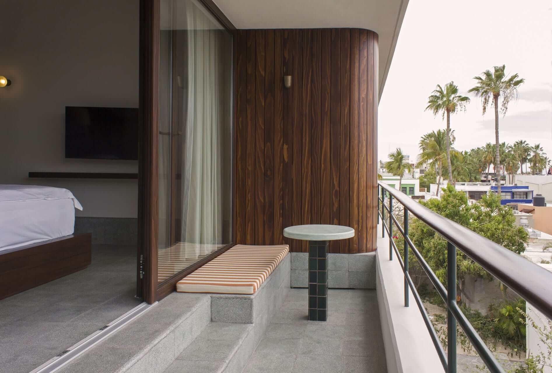 Baja Club, Mexico — Modern hotel room balcony with striped bench, wood paneling, coastal palm trees, and contemporary railing overlooking beachfront promenade.