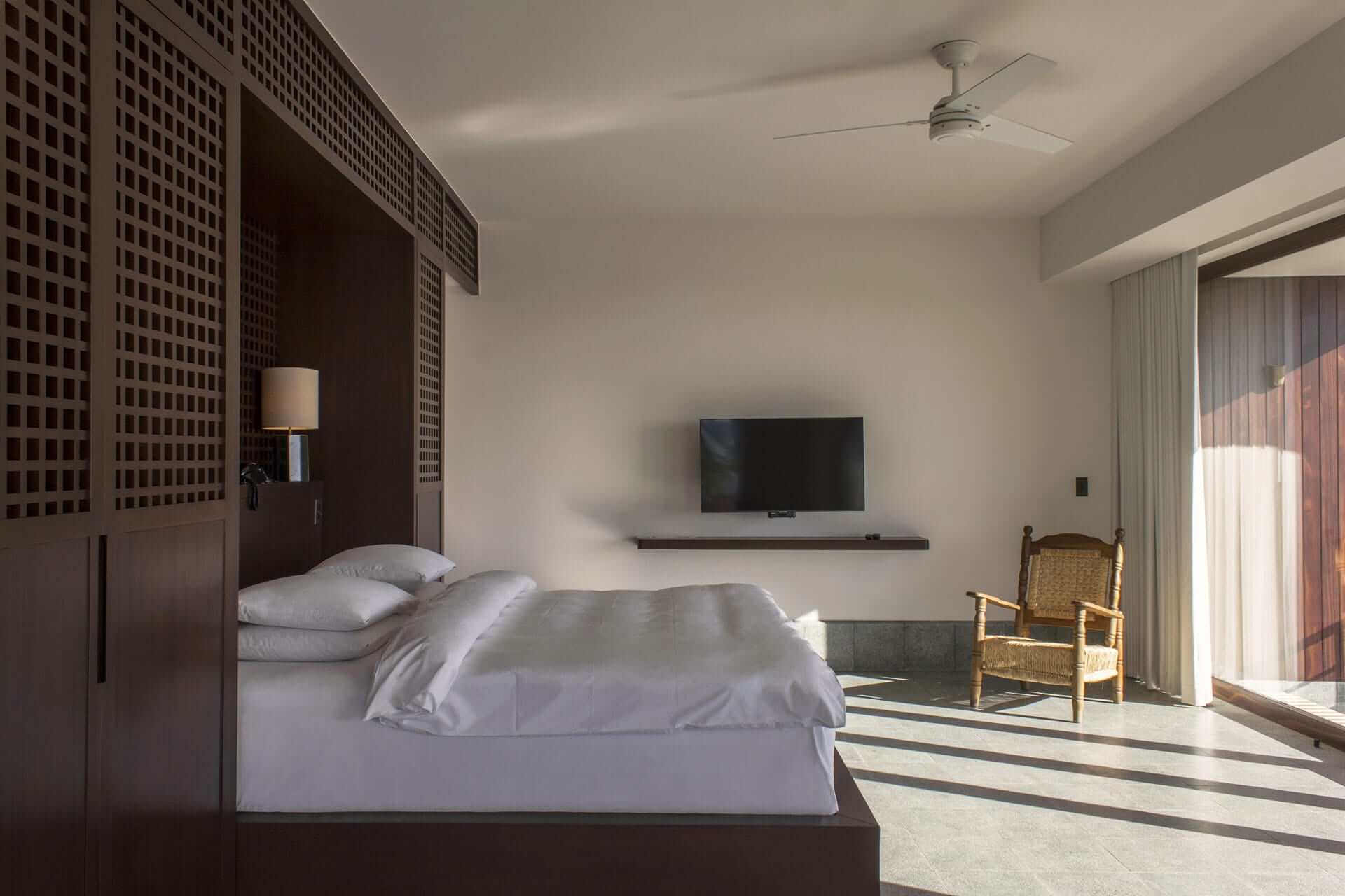 Baja Club, Mexico — Minimalist hotel bedroom with dark wood headboard wall, floating TV, woven chair, floor-to-ceiling windows, striped marble flooring, and soft natural light.