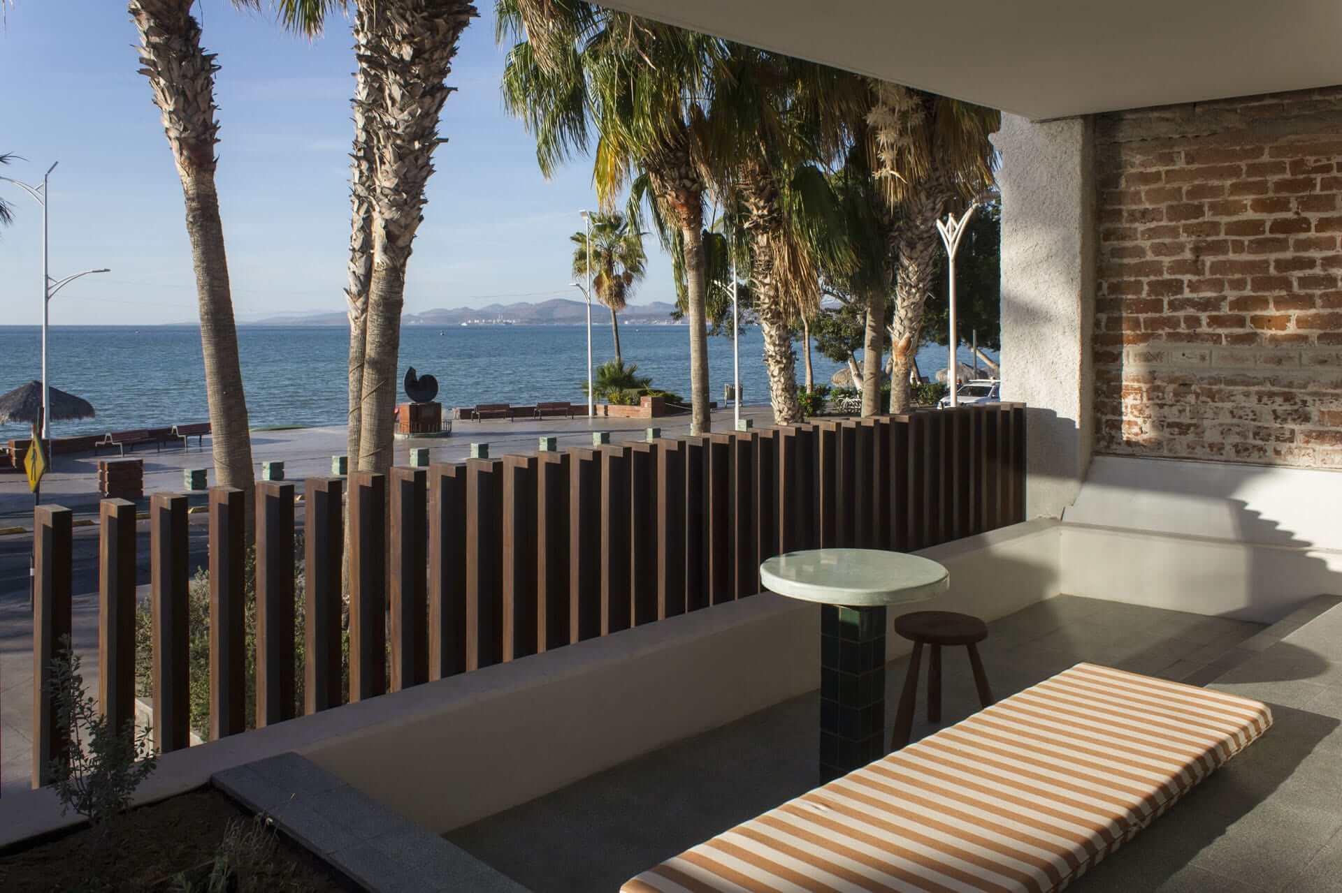 Baja Club, Mexico — Waterfront terrace with dark wood railing, striped lounge chair, round table, palm trees, and ocean view under natural light.