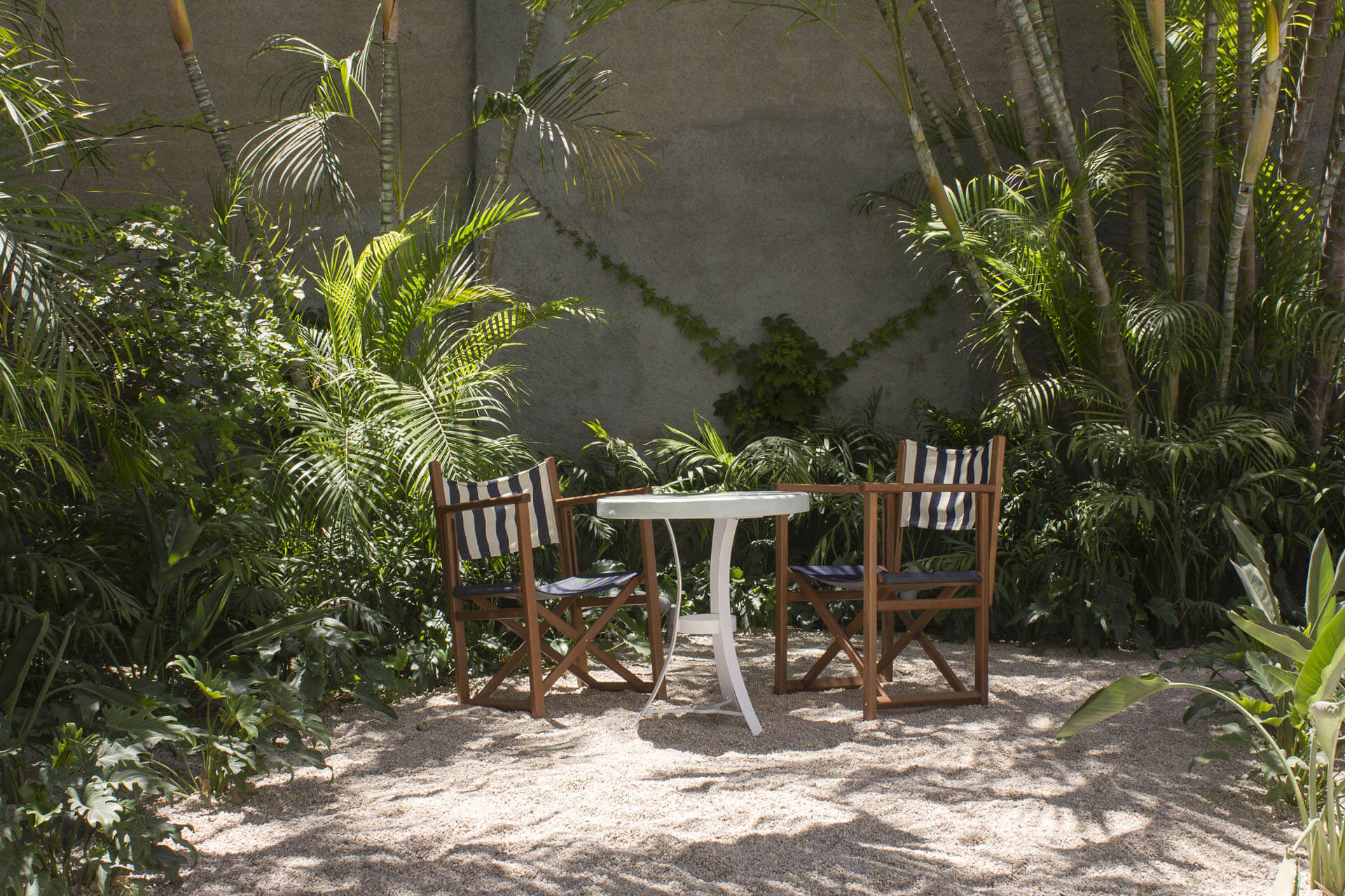 Baja Club, Mexico — Striped wooden chairs and white pedestal table nestled in sand courtyard surrounded by palm trees and concrete wall with ivy.
