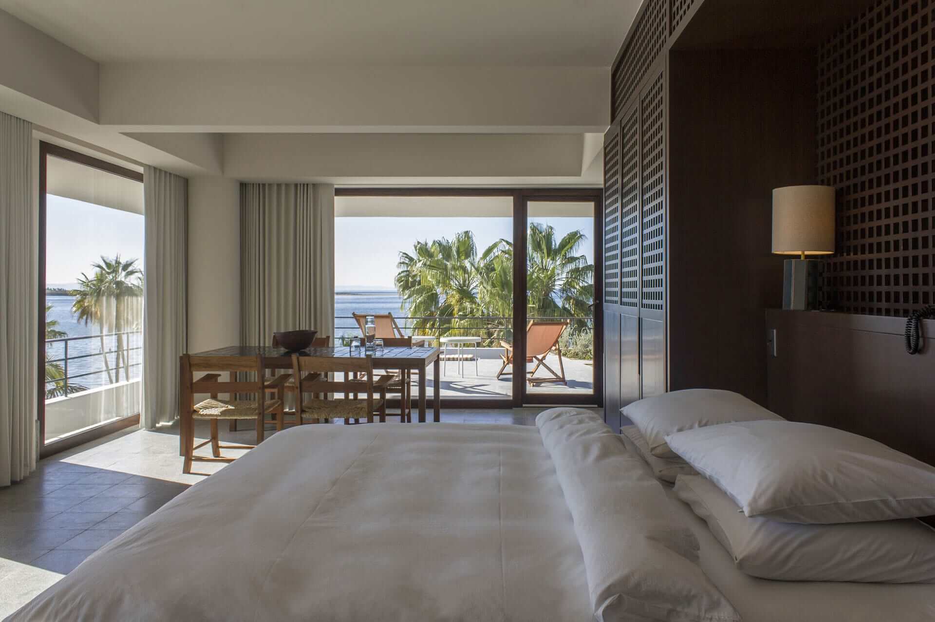 Baja Club, Mexico — Oceanfront hotel suite with dual beds, dark wood accent wall, floor-to-ceiling glass doors opening to sea-view terrace with palm trees and lounge chairs.