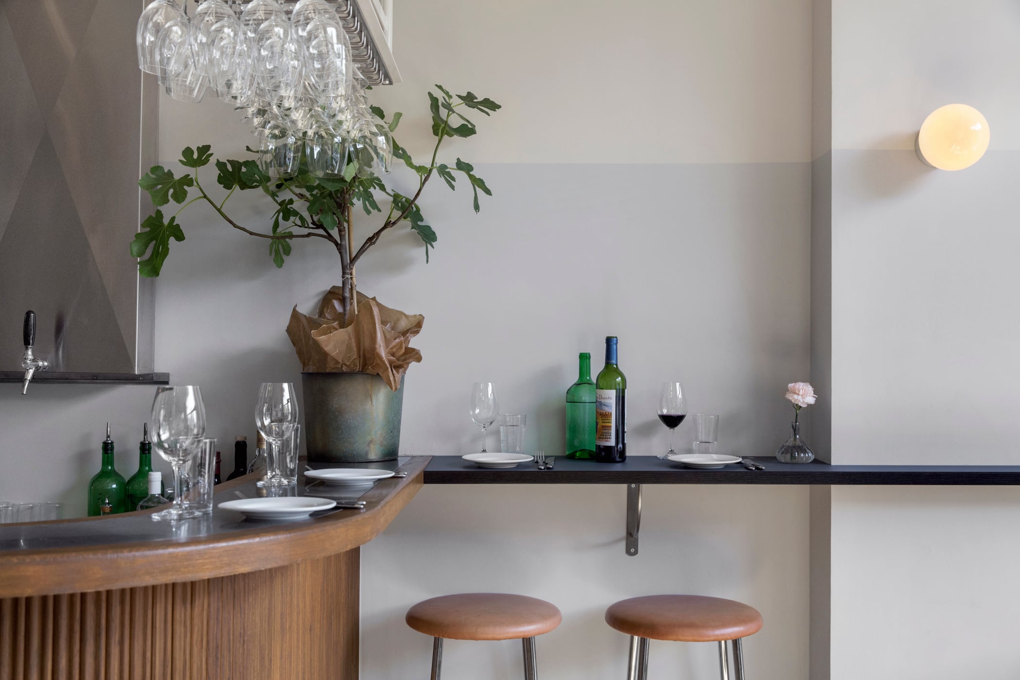 Bar Agrikultur, Europe — Minimalist bar counter with curved wooden base, potted plant under crystal pendant, wine bottles and place settings on floating shelf, leather stools, warm ambient lighting.