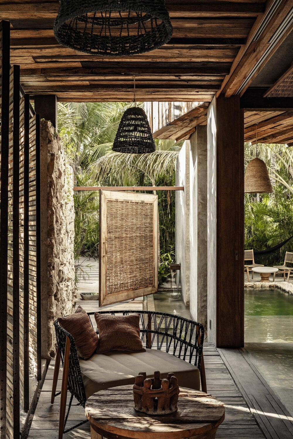 Be Tulum, Mexico — Black woven pendant lamp and rattan daybed on wooden deck overlooking tropical lagoon with palm trees and stone walls.