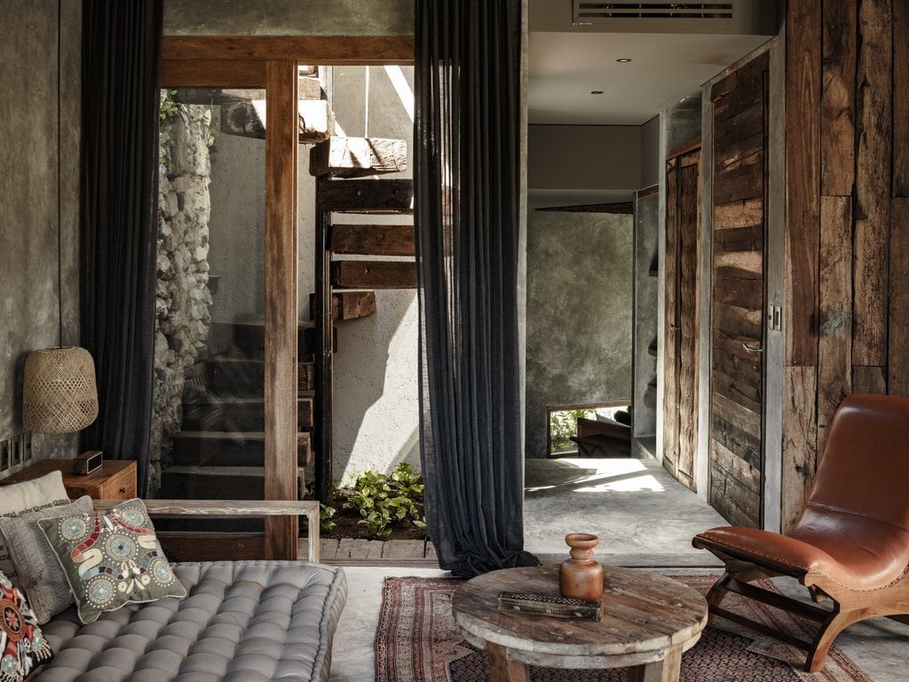 A sunlit passage at Be Tulum frames weathered wood doors, concrete walls, and mid-century seating beneath dark curtains.