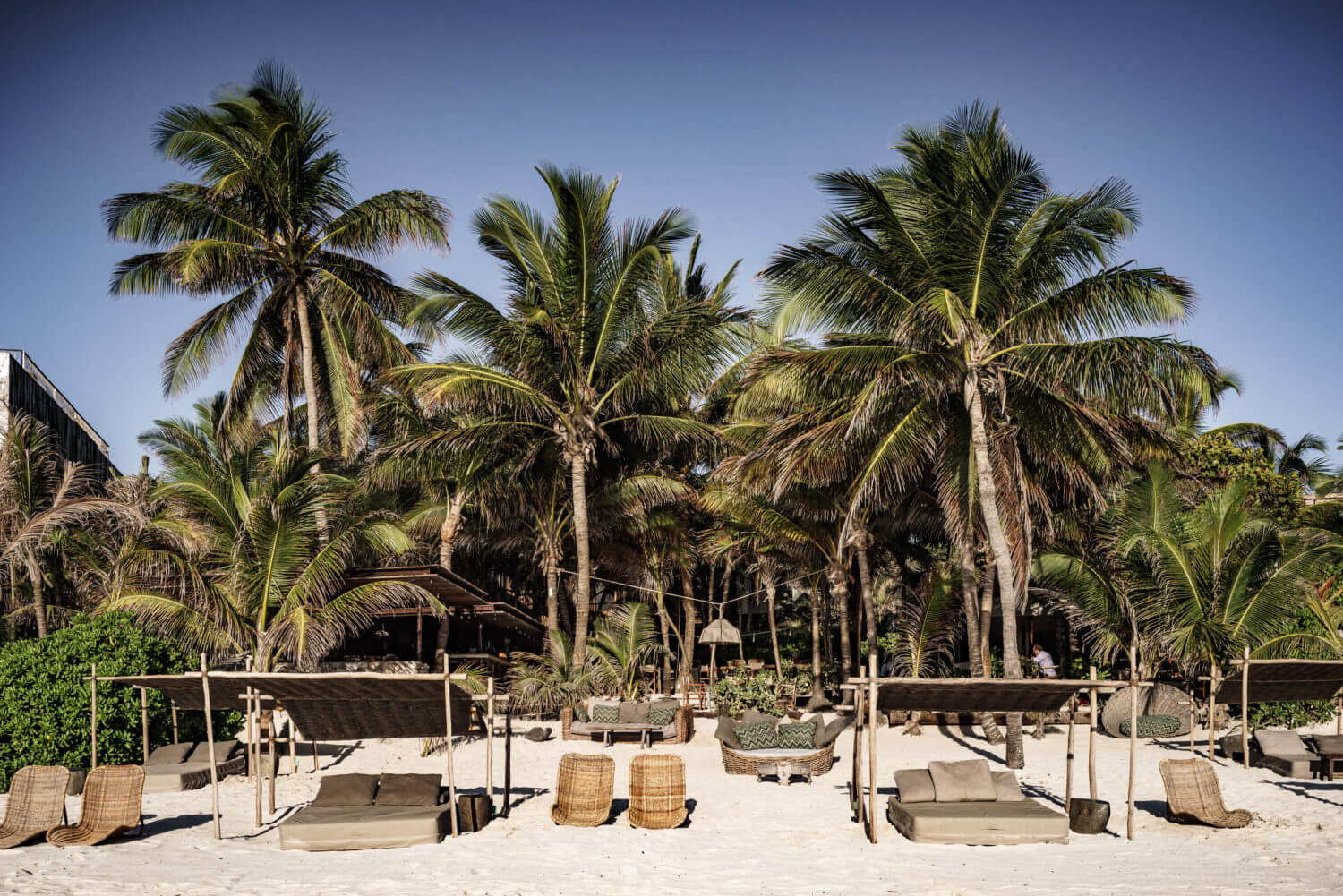 Be Tulum, Tulum, Mexico — Beach club with woven cabanas, palm trees, and sandy lounge area under clear sky at luxury resort.