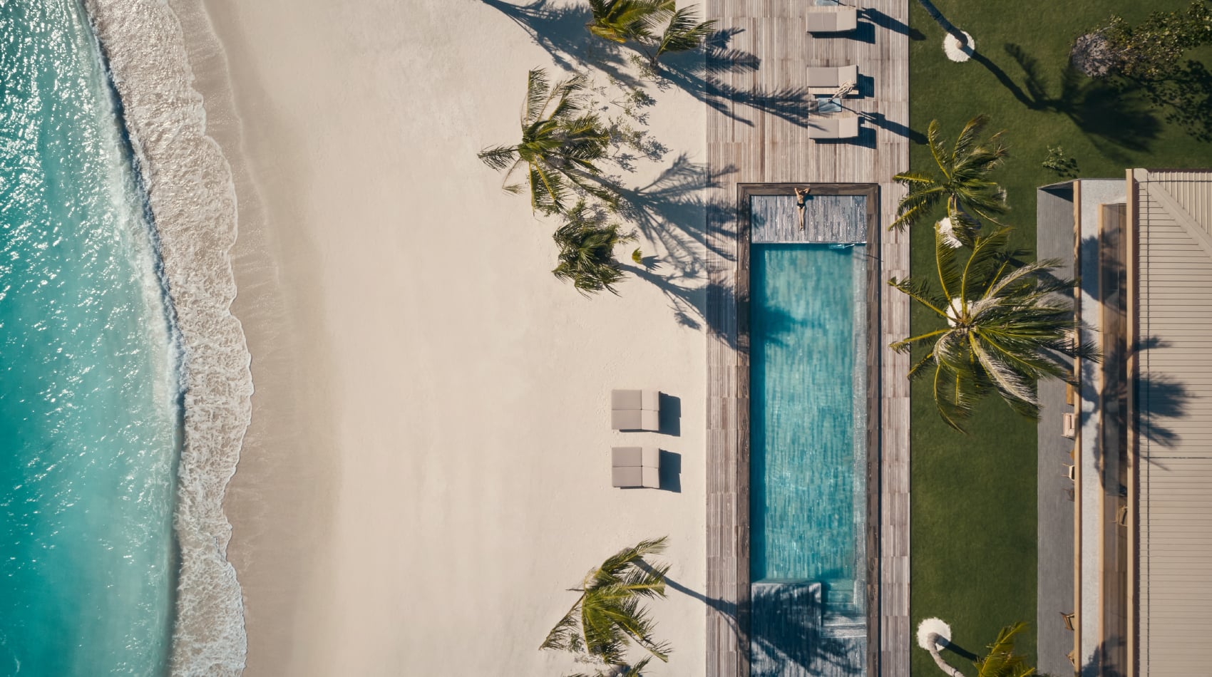 Patina Maldives, Asia — Aerial view of beachfront resort with turquoise infinity pool, wooden deck, white sand, palm trees, and modern architecture overlooking tropical ocean.