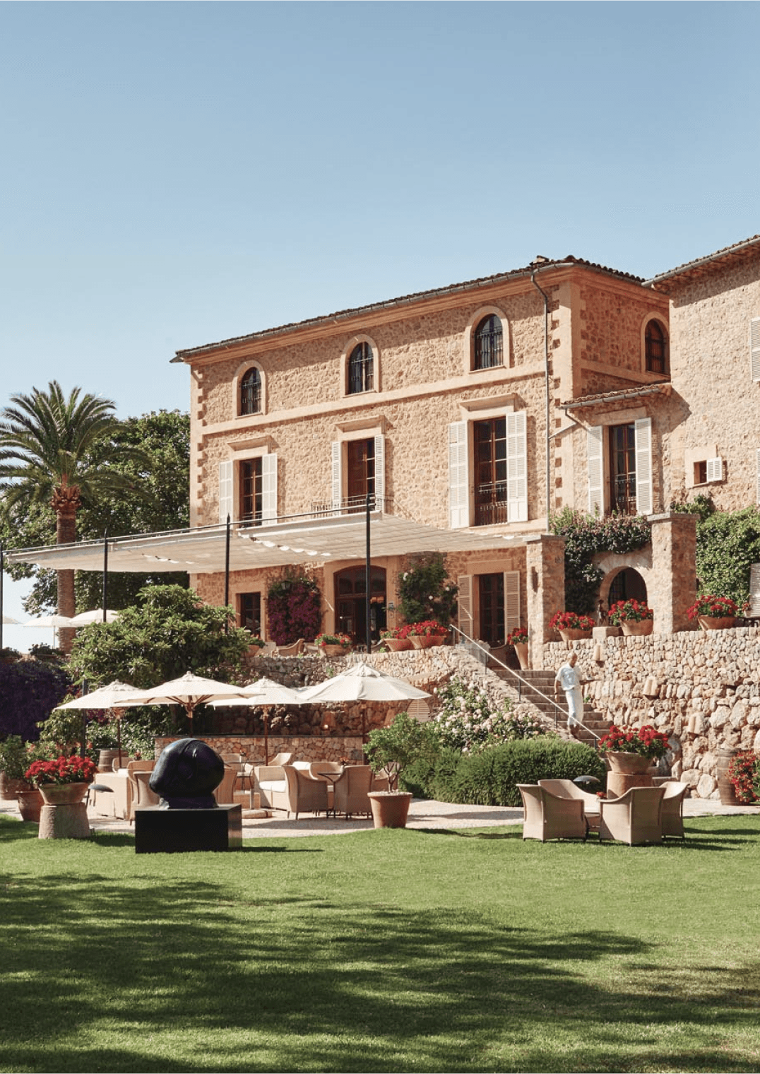 Belmond La Residencia, Balearic Islands — Stone villa with terracotta trim, manicured lawn, umbrellas, and Mediterranean garden in golden afternoon light.