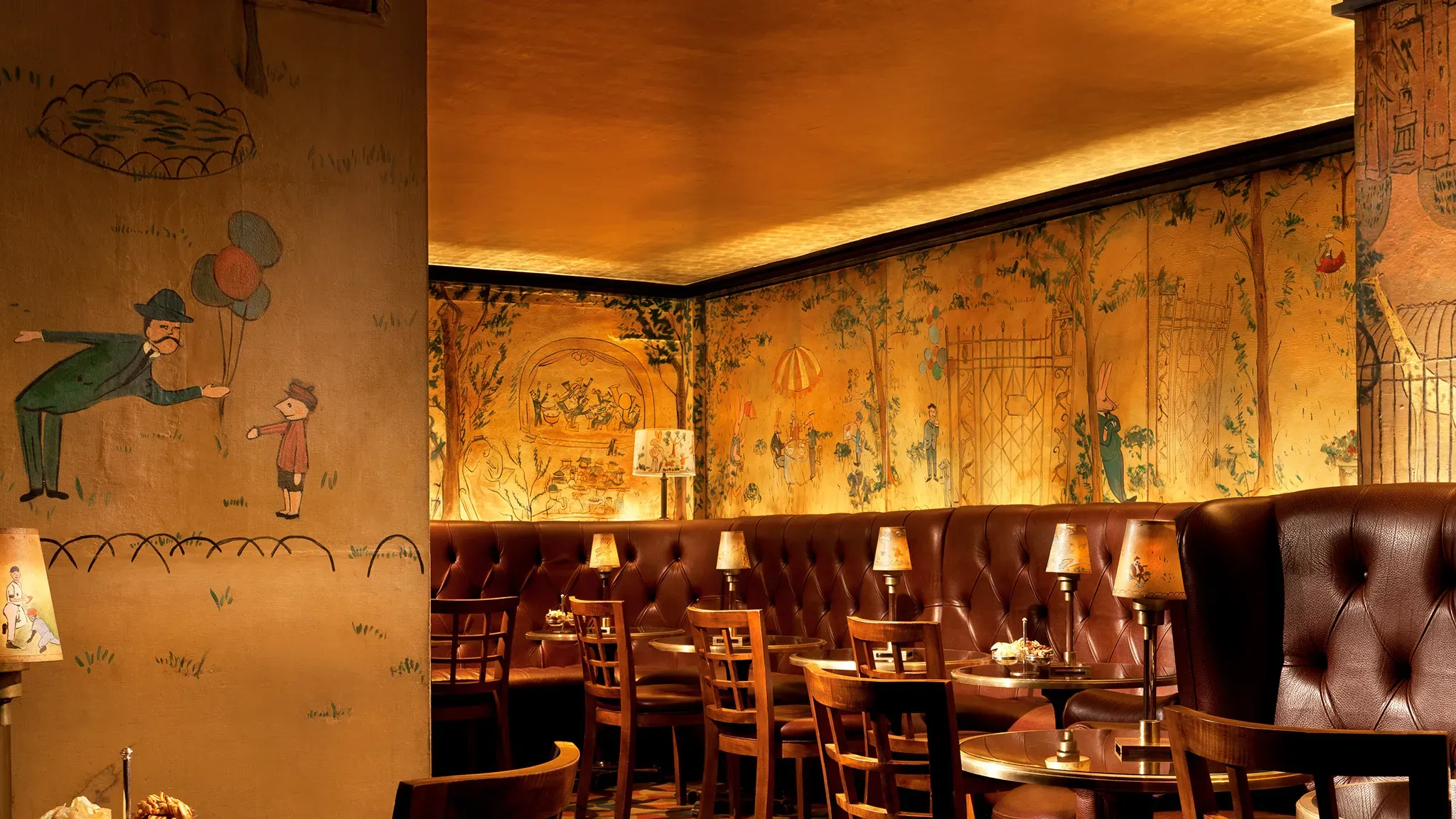 Bemelmans Bar, New York, United States — Warm-lit dining room with hand-painted murals, burgundy leather banquette seating, brass table lamps, and wood chairs under glowing ceiling.