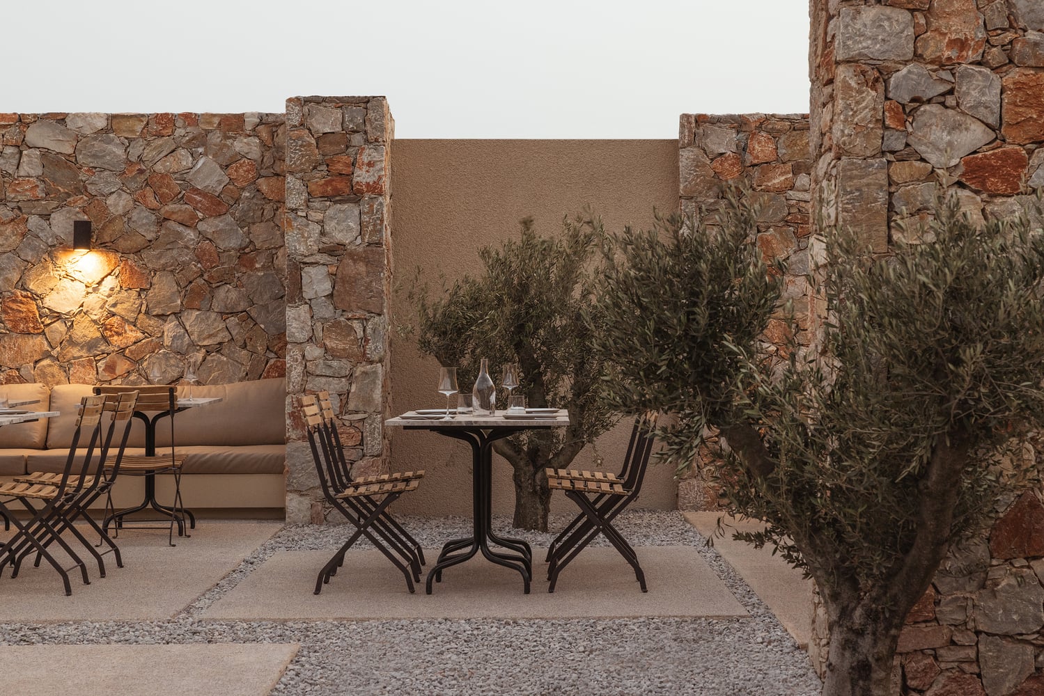Gundari, Greece — Stone courtyard with rustic ochre walls, olive trees, and candlelit dining table at dusk.