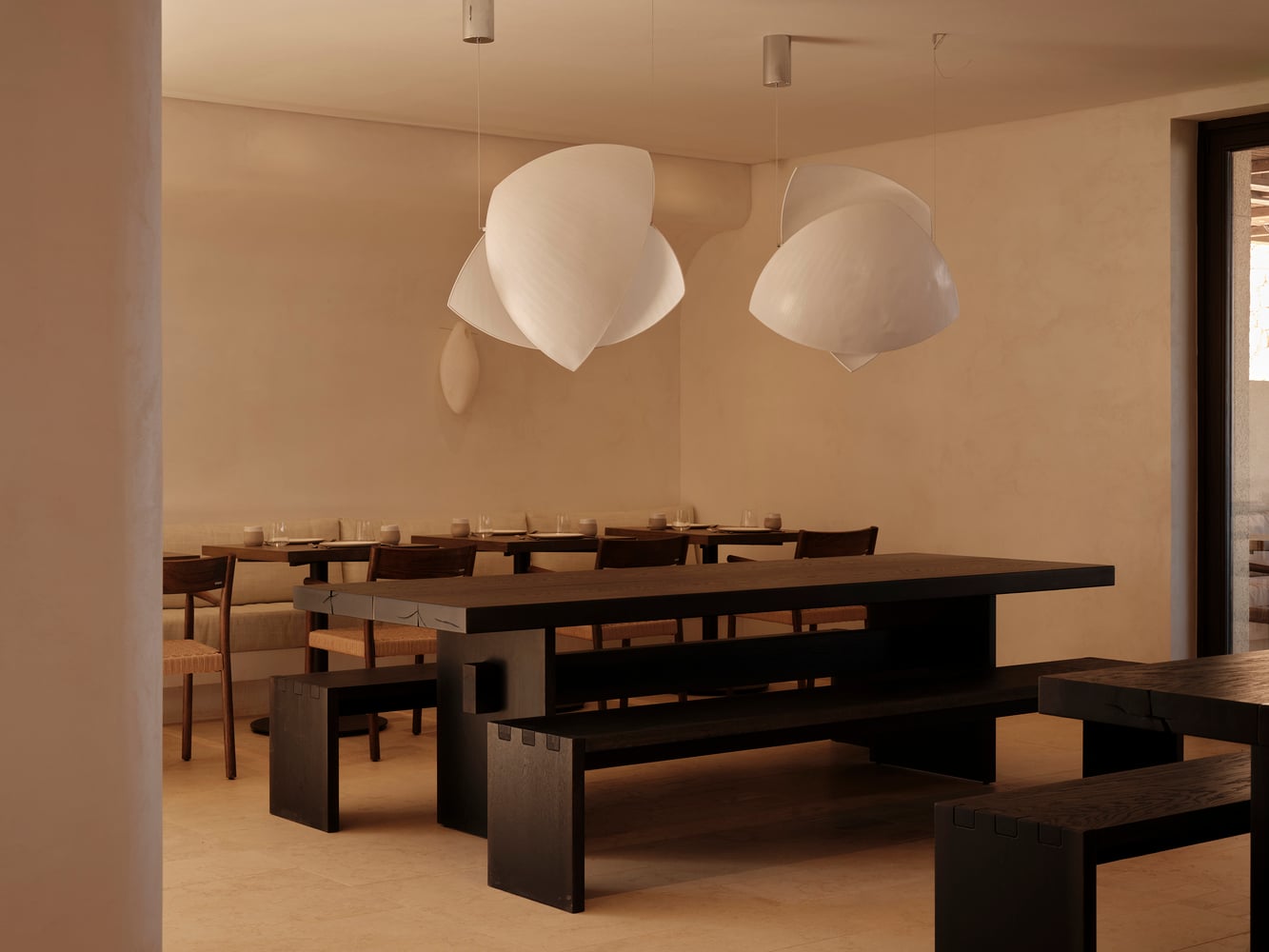 Gundari, Greece — Minimalist dining room with dark wood tables, cream pendant lights, beige walls, and natural light from clerestory windows.