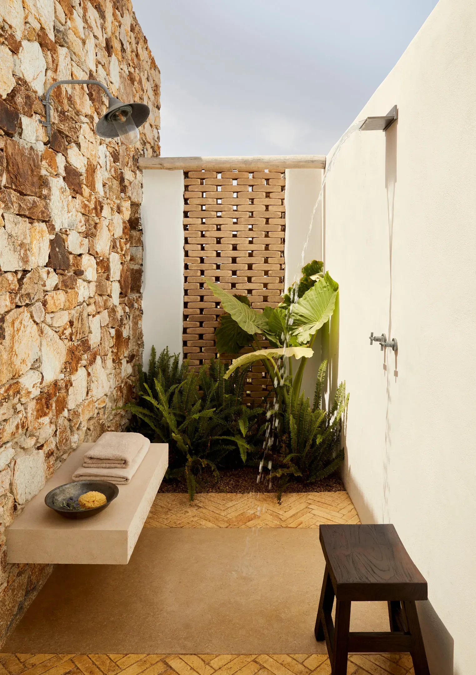 The Rooster, Europe — Outdoor shower courtyard with stone wall, wooden lattice screen, potted plants, concrete bench, and herringbone tile flooring.