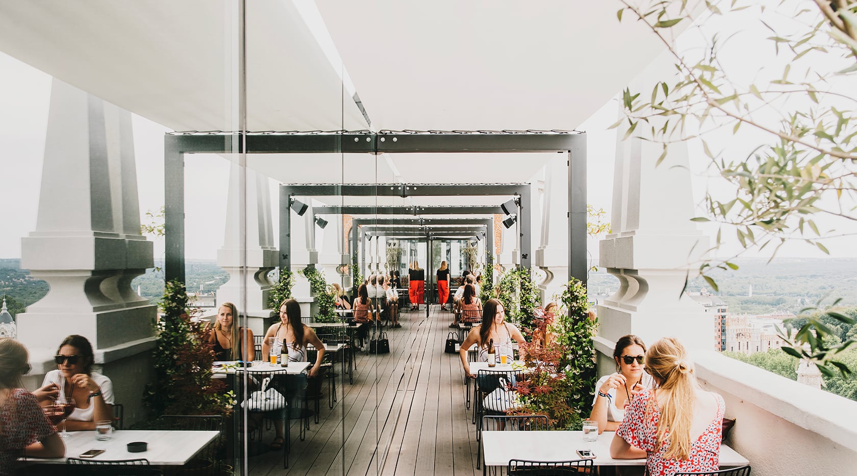 Nice To Meet You Restaurant & Lounge, Europe — Elevated terrace with glass partitions, wooden decking, climbing greenery, and panoramic valley views, guests dining at white tables under industrial track lighting.
