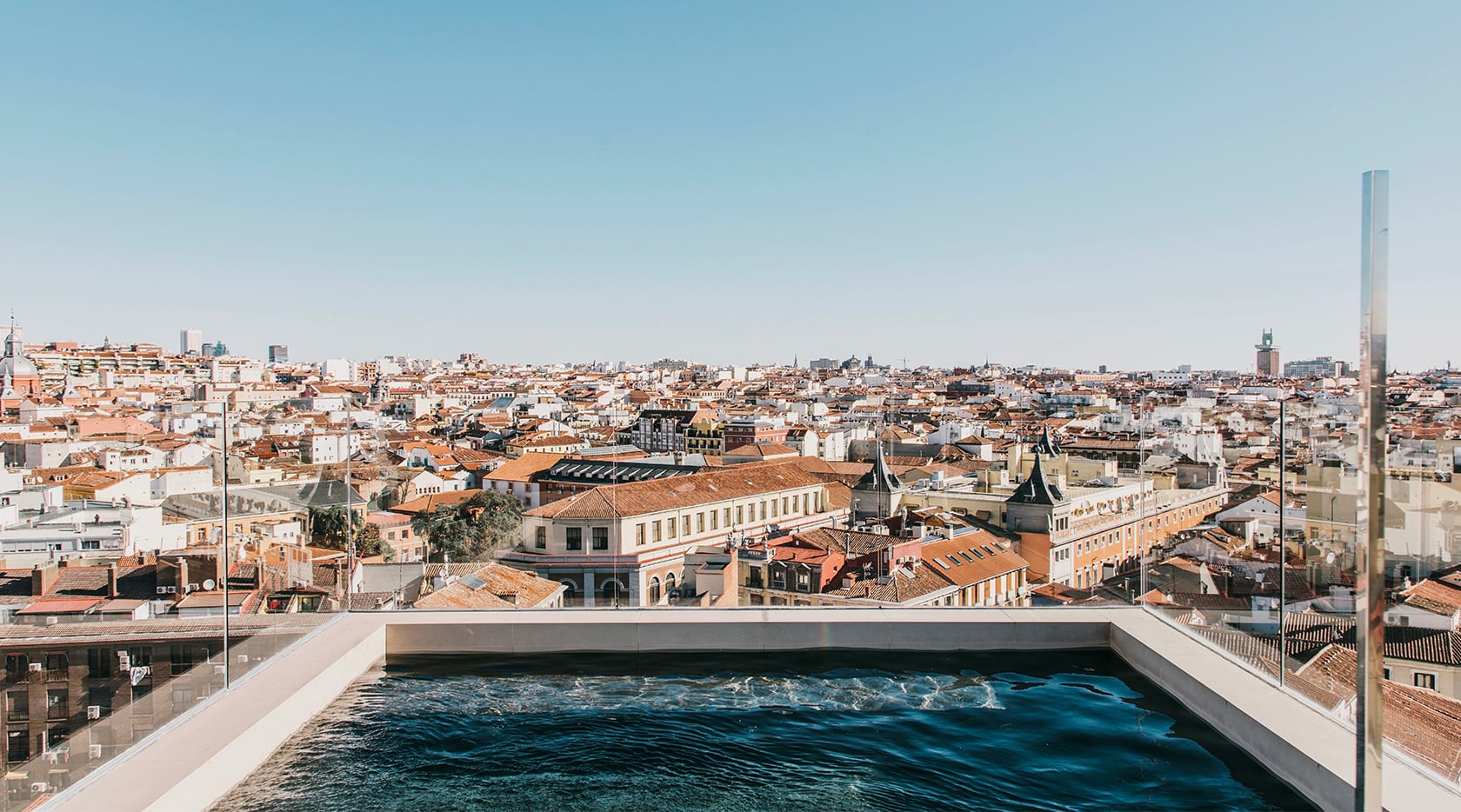 Rooftop pool terrace overlooking European city with terracotta-tiled buildings, modern minimalist concrete deck, clear blue water, and urban skyline at golden hour.