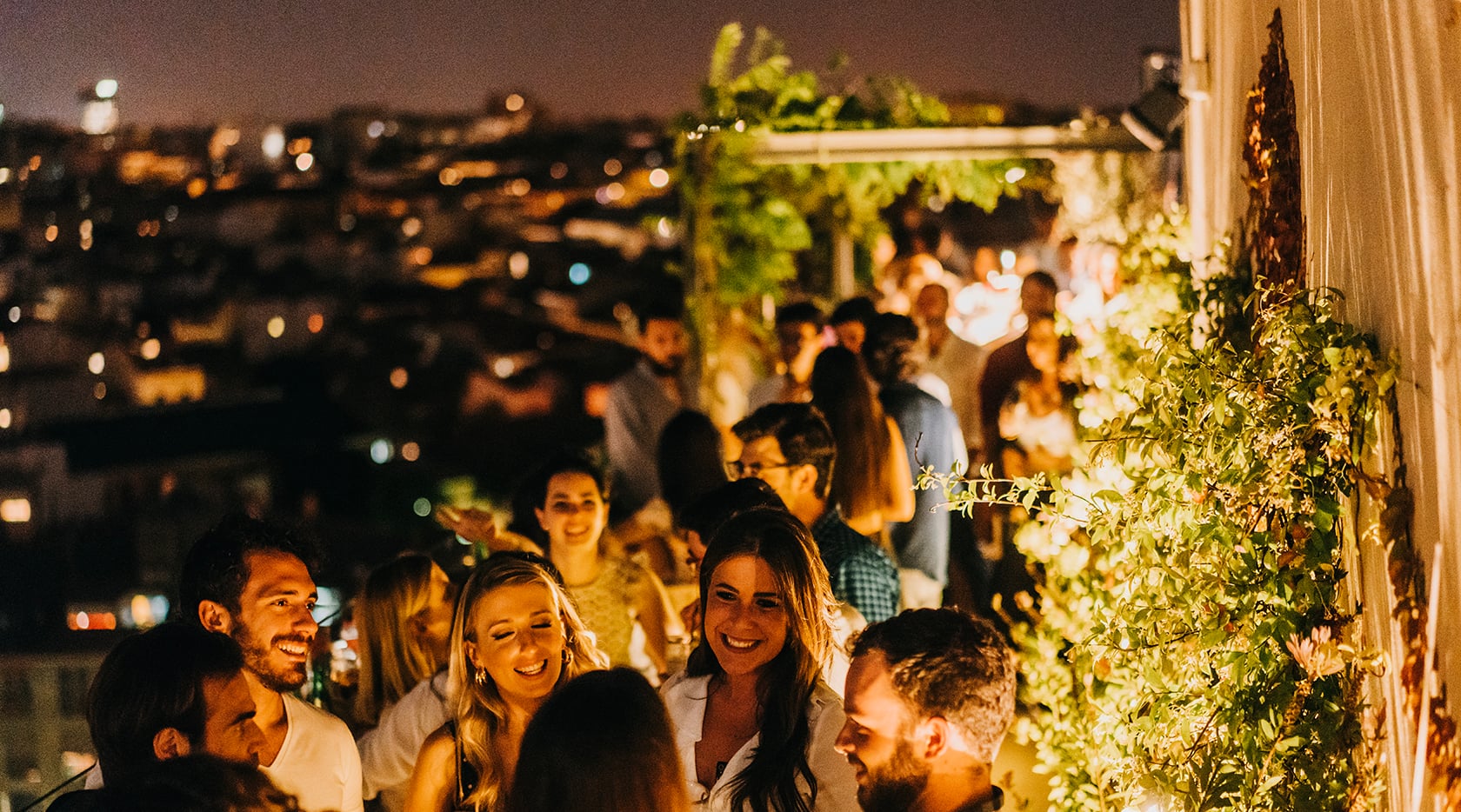 Dear Hotel, Europe — Rooftop terrace at dusk with ivy-draped columns, warm lighting, city skyline, and guests mingling under evening glow.