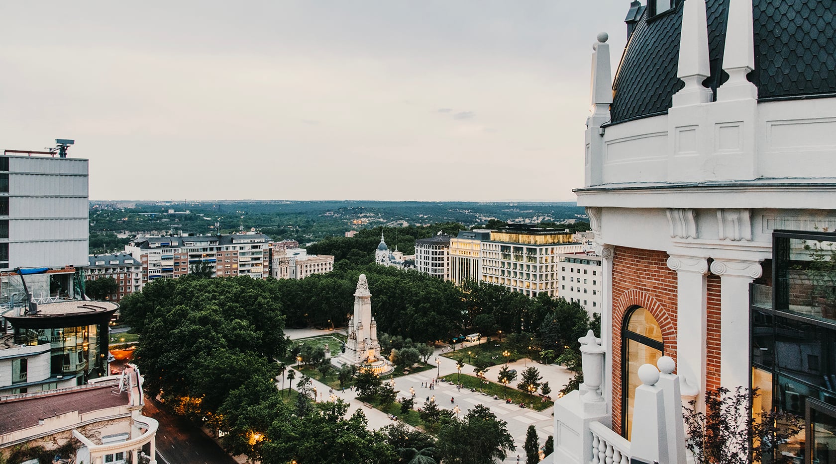 Dear Hotel, Europe — Aerial view of classical monument in verdant plaza surrounded by belle époque architecture, green canopy, and distant cityscape under overcast sky.
