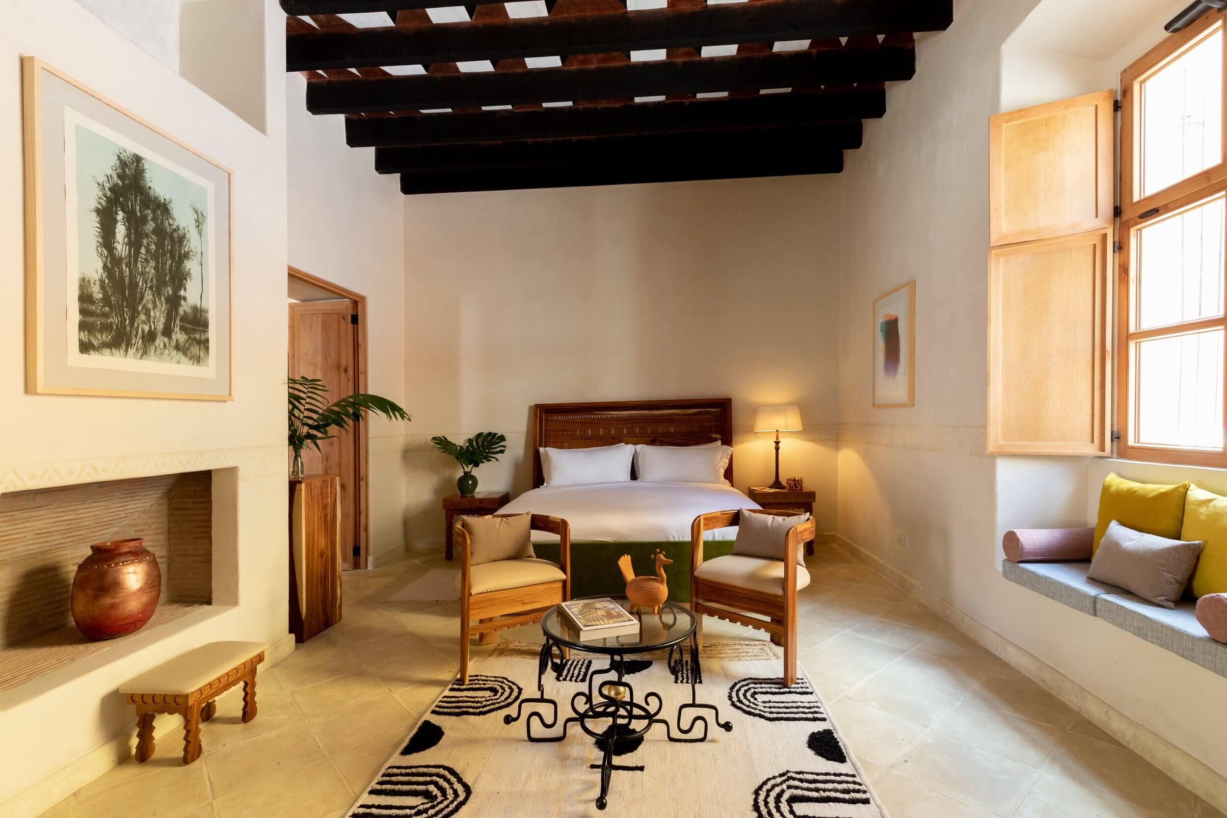 La Valise San Miguel, Mexico — Minimalist bedroom with dark wooden beams, terracotta pottery, geometric patterned rug, and natural light from large windows.