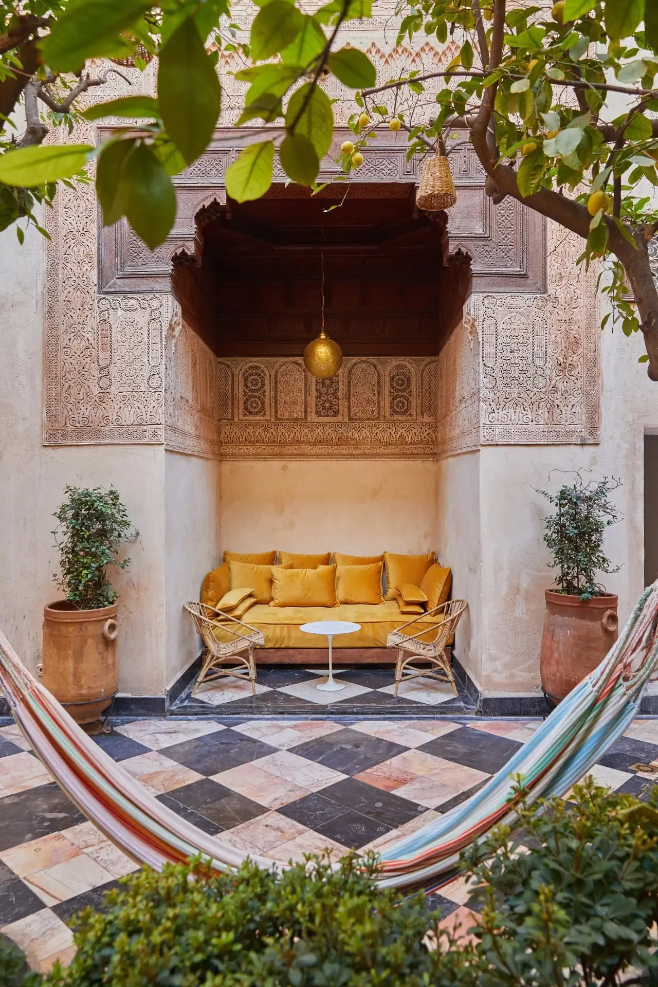 El Fenn, Marrakech, Morocco — Ornate carved cedarwood alcove with golden cushioned daybed, geometric tile flooring, potted plants, and hanging brass lantern in Moroccan riad courtyard.