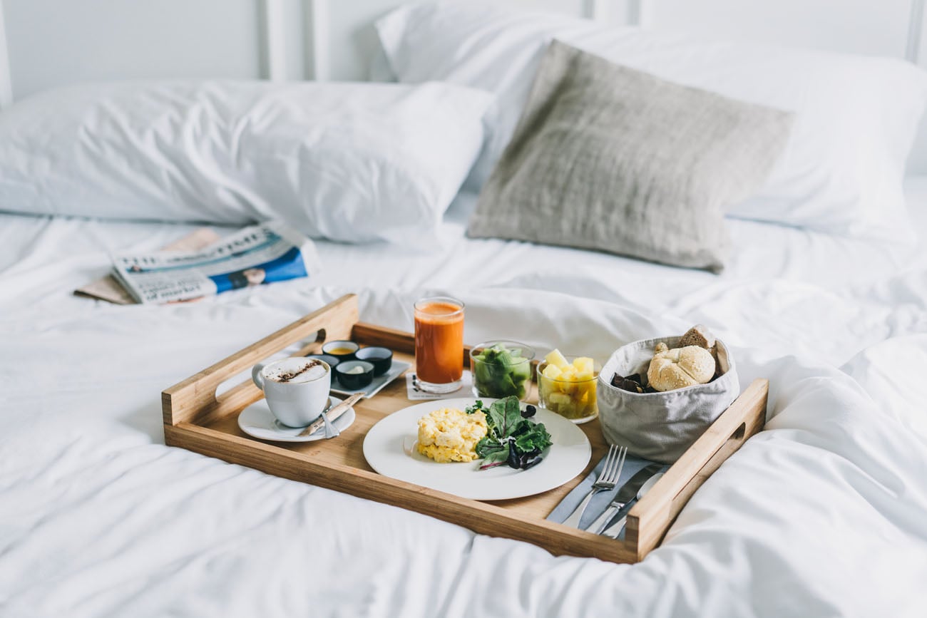 Dear Hotel, Europe — Wooden breakfast tray with scrambled eggs, greens, coffee, orange juice, and bread basket on white linens and pillows.
