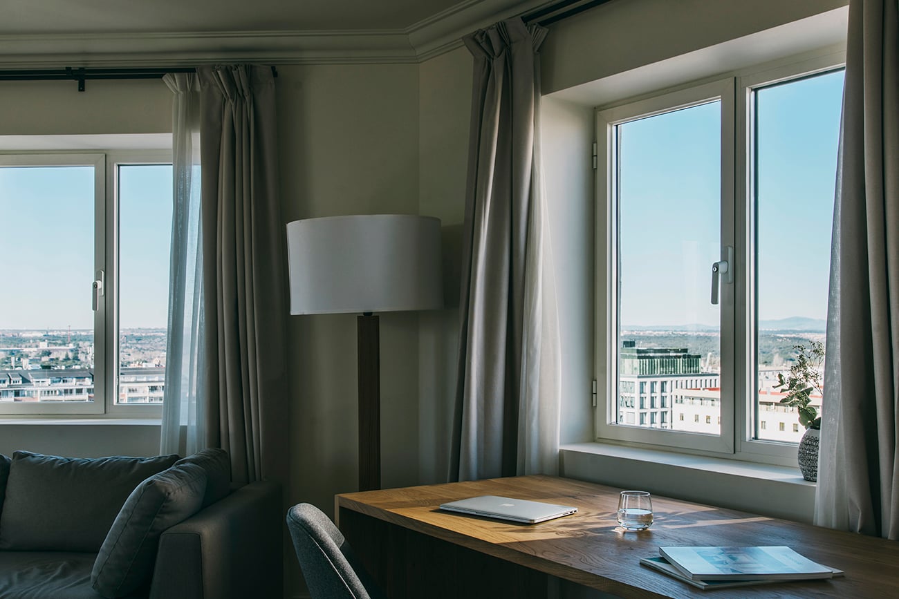 Dear Hotel, Europe — Modern hotel suite with floor-to-ceiling windows overlooking cityscape, minimalist furnishings, natural light, contemporary floor lamp, wooden desk.