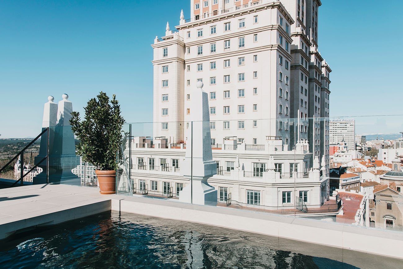 Dear Hotel, Europe — Modernist rooftop terrace with infinity pool, cream limestone facade, potted greenery, coastal cityscape, clear blue sky, afternoon light.