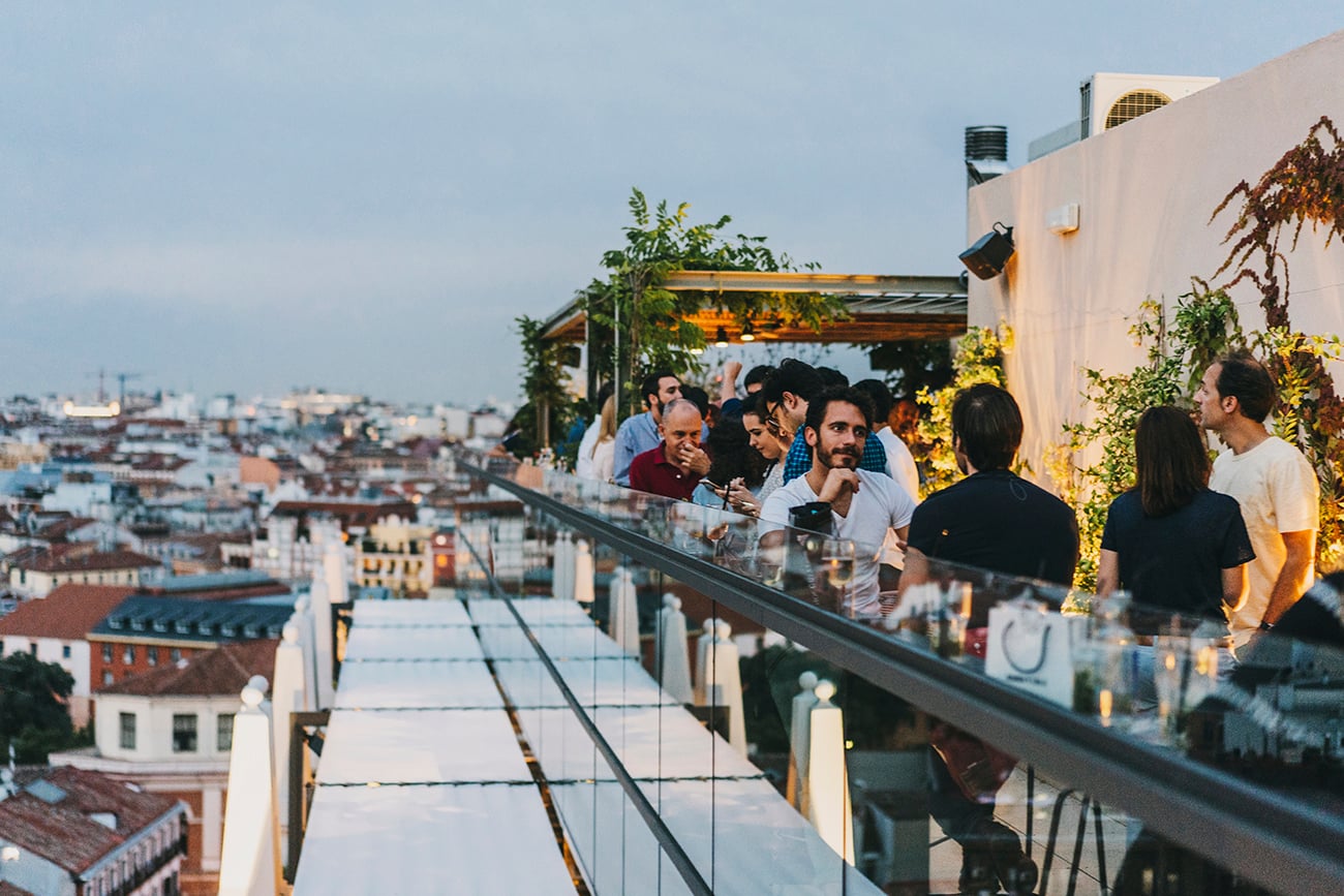 Dear Hotel, Europe — Rooftop terrace gathering at dusk with city skyline views, minimalist white architecture, string lights, and lush greenery.
