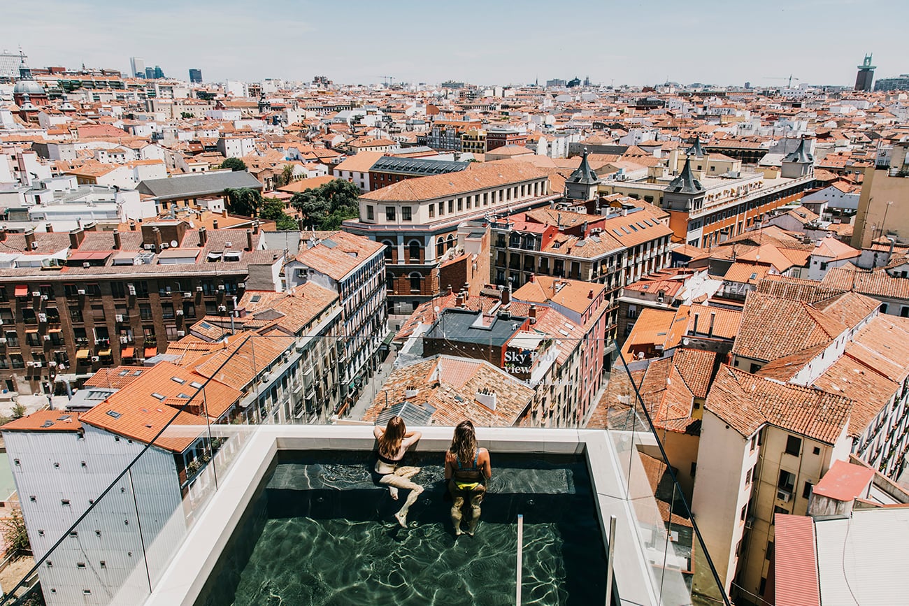 Dear Hotel, Madrid, Spain — Two guests swimming in rooftop pool overlooking terracotta-tiled city skyline and historic architecture in afternoon light.