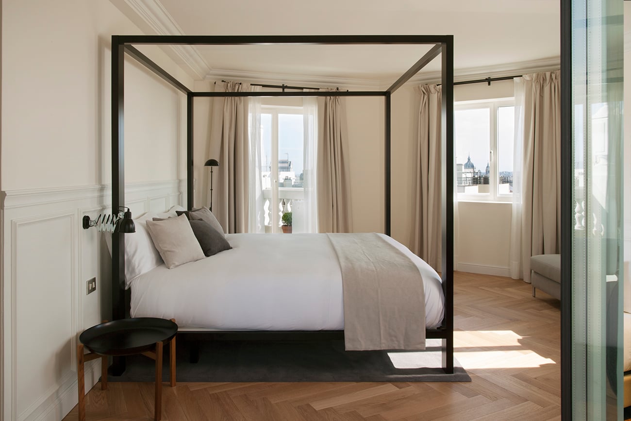 Dear Hotel, Europe — Black four-poster bed with white linens in minimalist bedroom with floor-to-ceiling windows overlooking city skyline, natural light, herringbone flooring.