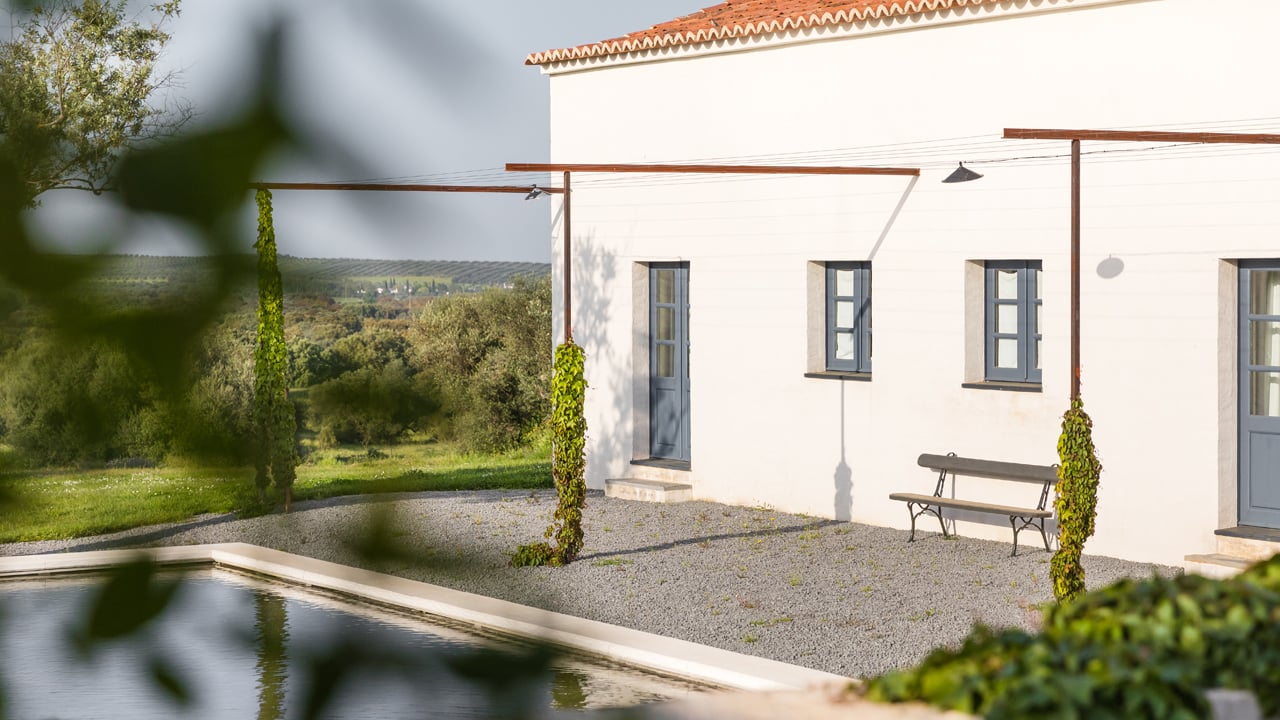São Lourenço do Barrocal, Portugal — Modernist whitewashed residence with terracotta roof, ivy-clad pergola, gravel courtyard, bench seating, and pastoral landscape views.
