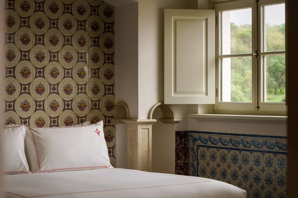 Vermelho, Europe — Bedroom with ornate patterned headboard, white linens with red trim, framed mirror, and large windows overlooking green countryside.