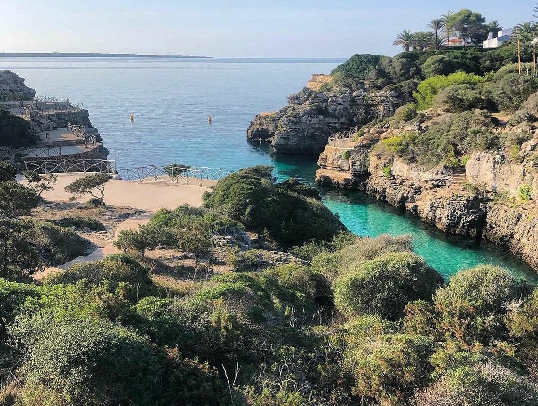 Cala en Brut, Balearic Islands, Spain — turquoise cove nestled between limestone cliffs, Mediterranean vegetation, sandy beach with wooden boardwalk, calm azure waters.
