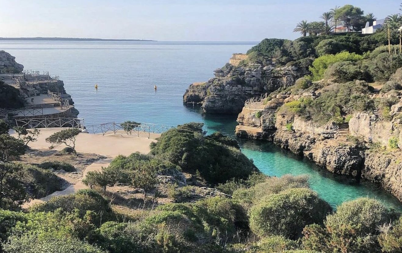 Cala en Brut, Balearic Islands, Spain — turquoise cove nestled between limestone cliffs, Mediterranean vegetation, sandy beach with wooden railings and sun loungers.