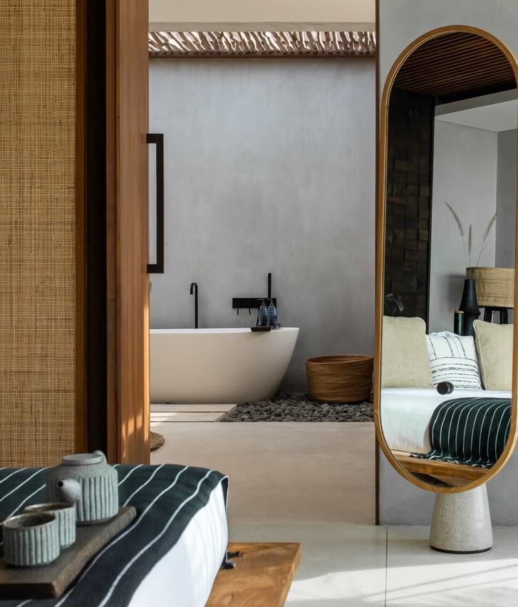 Cap Karoso, Asia — Minimalist ensuite with freestanding stone bathtub, woven rattan screens, brass mirror, and striped bedding in neutral tones.