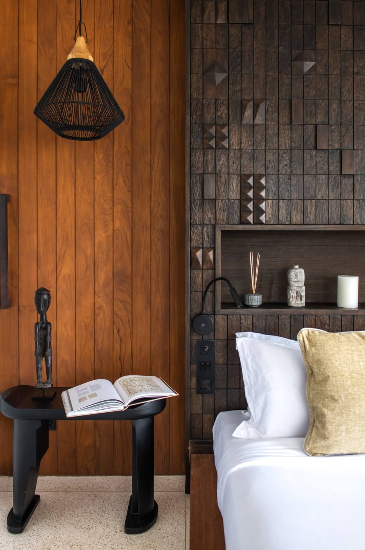 Cap Karoso, Asia — Minimalist bedroom with warm wooden wall paneling, woven pendant light, dark geometric tile accent, floating shelf with candles, and layered white bedding with mustard cushion.