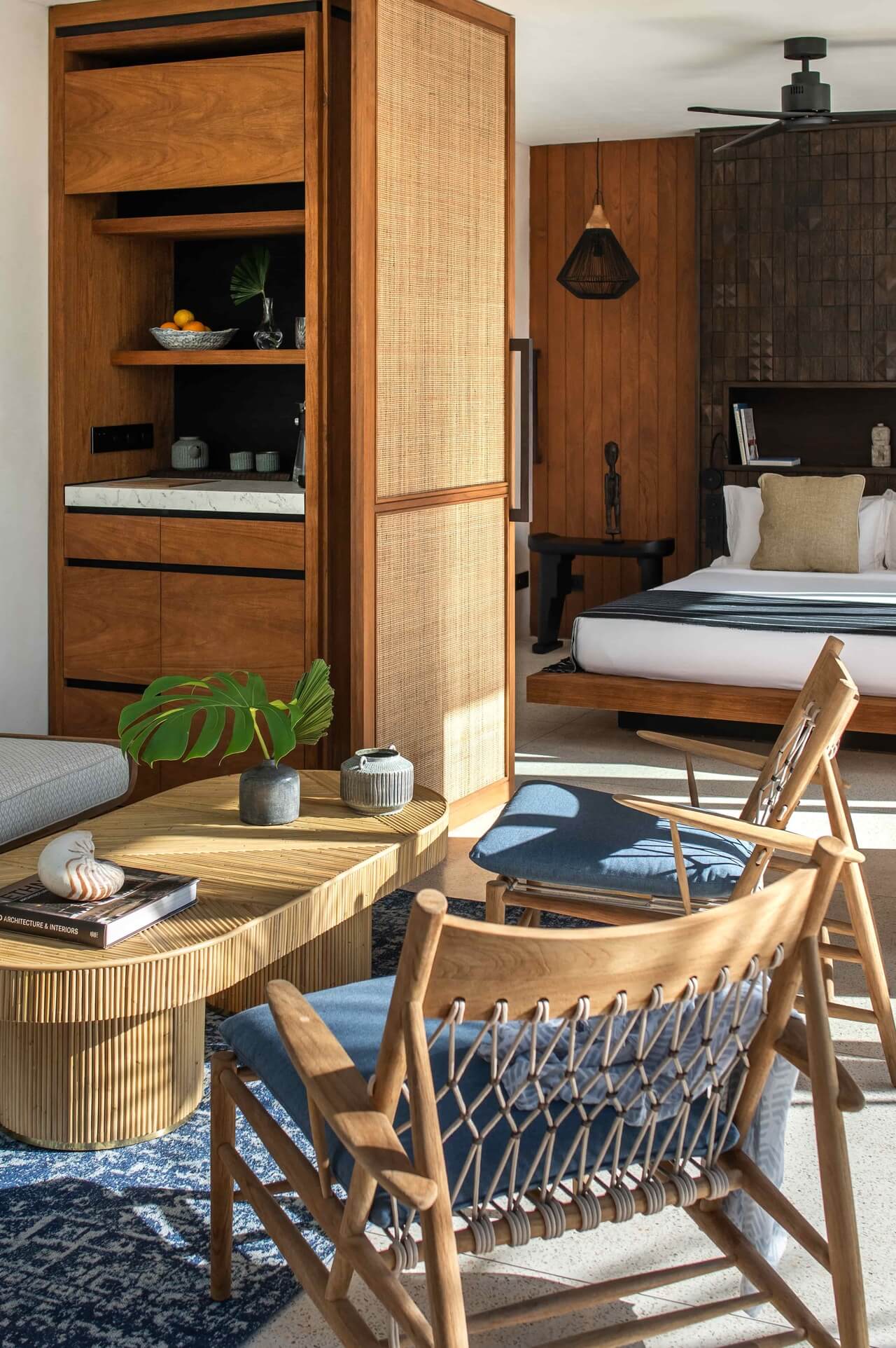 Cap Karoso, Asia — Tropical resort bedroom with teak wood furnishings, woven rattan room divider, mid-century modern chairs, and natural light filtering through wooden slat ceiling.