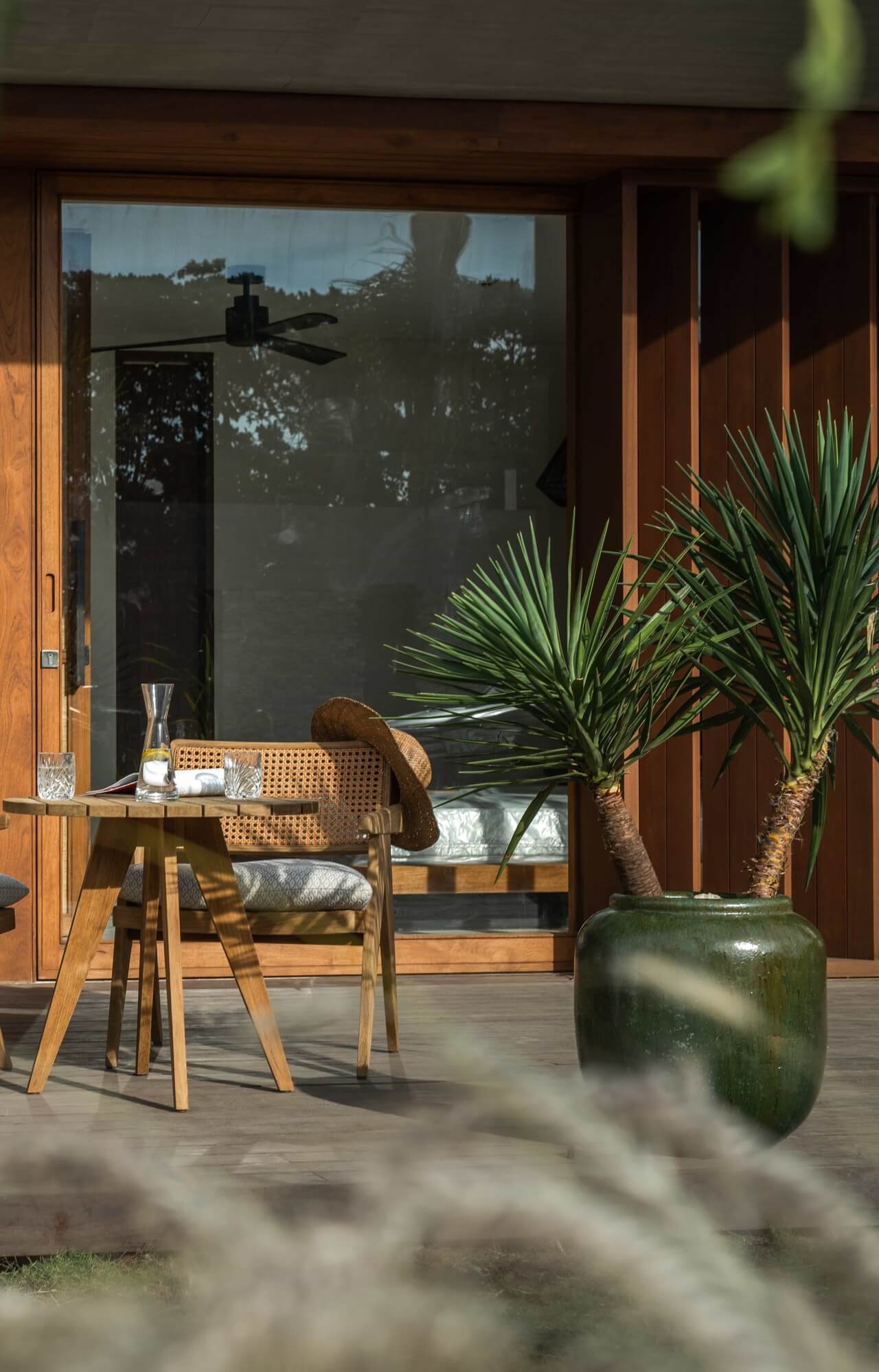 Cap Karoso, Asia — Wooden deck with rattan chair, potted palm, and glass doors overlooking forested landscape, morning light casting shadows.