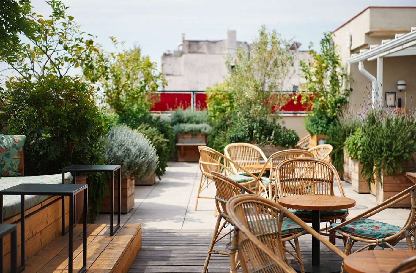Casa Bonay, Barcelona, Spain — Rooftop terrace with woven rattan chairs, potted herbs, wooden deck, and city views framed by greenery.