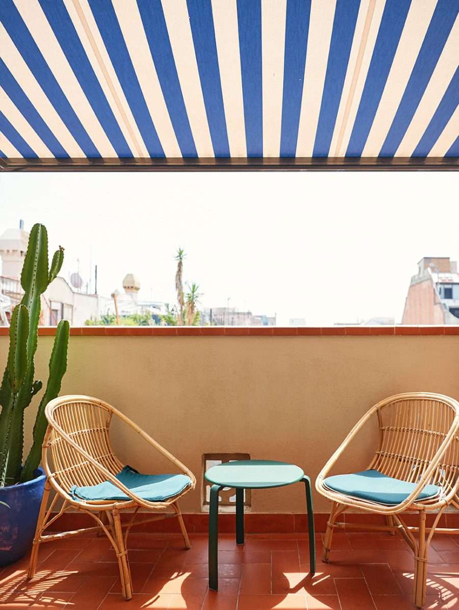 Casa Bonay, Europe — Striped awning shades rattan lounge chairs with blue cushions on terracotta terrace overlooking cityscape with cacti and architectural details.