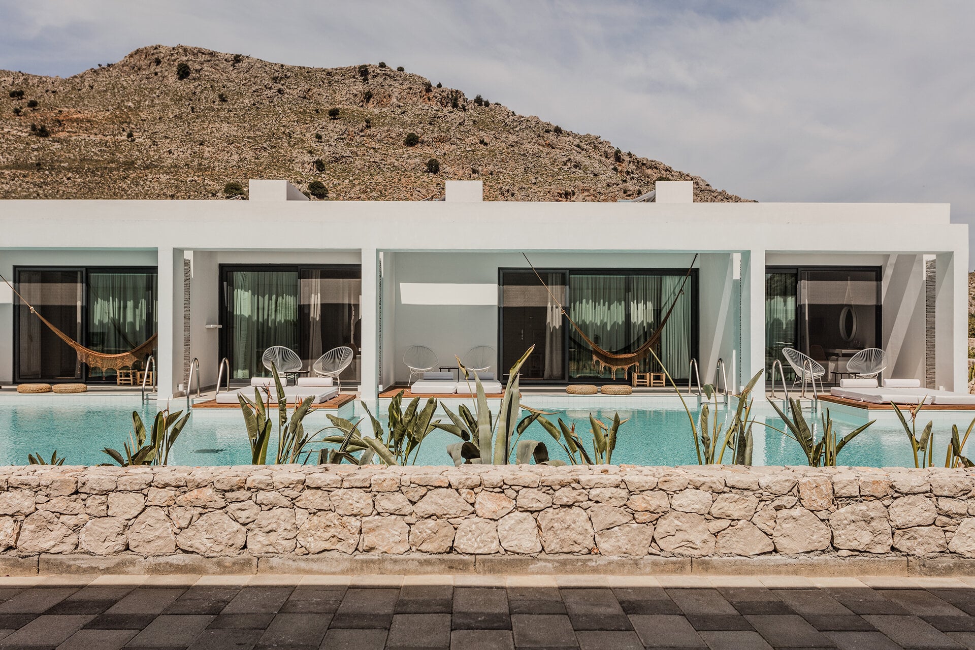 Casa Cook Rhodes, Greece — Modern whitewashed poolside suites with green-tinted glass, woven loungers, and stone terrace overlooking turquoise water against rocky hillside.