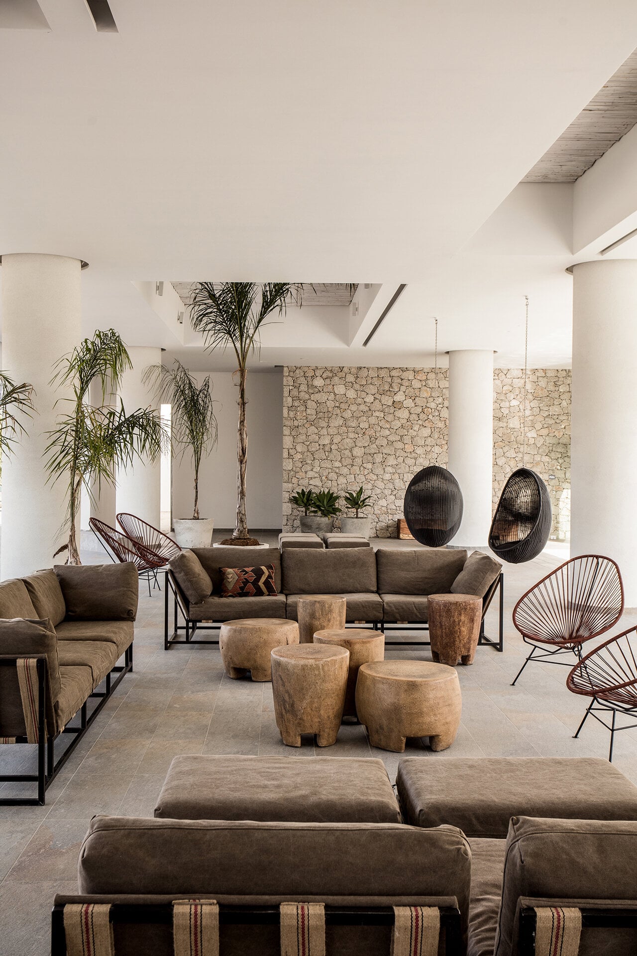 Casa Cook Rhodes, Greece — Minimalist lounge with natural stone walls, earth-toned upholstered seating, wooden sculptural stools, hanging rattan chairs, and tall potted palms beneath coffered ceiling.