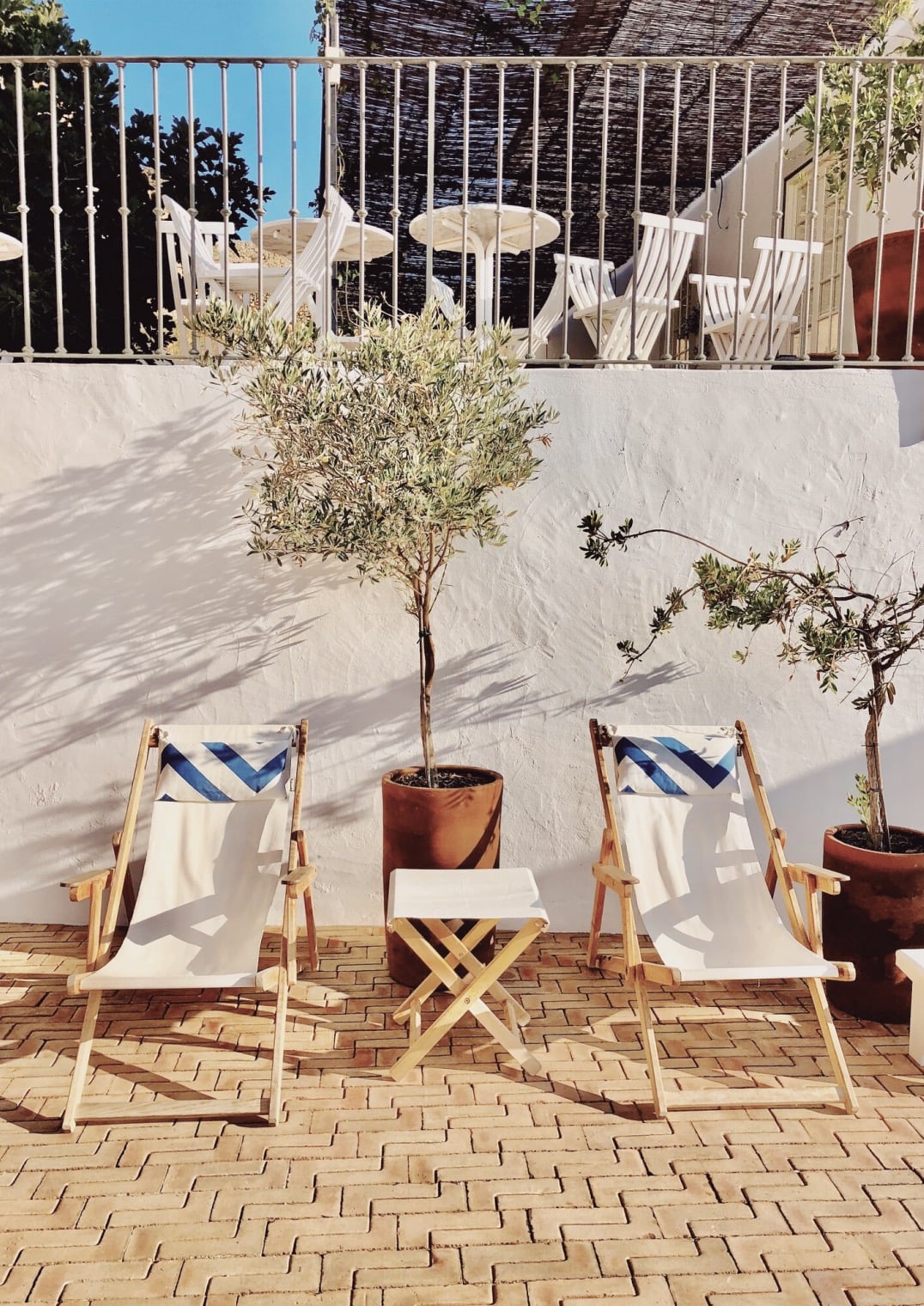 Casa Mãe, Europe — Striped deck chairs and potted olive trees on terracotta brick patio beneath white-washed walls and metal railings, Mediterranean courtyard design.
