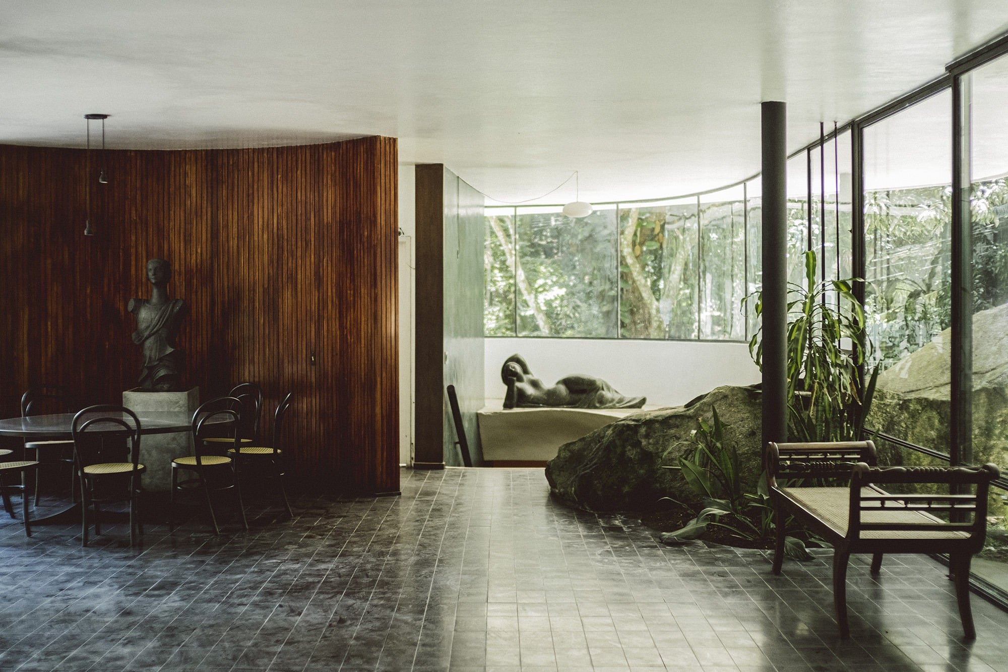 Oscar Niemeyer Casa das Canoas, Rio de Janeiro, Brazil — Modernist interior with wood-slat walls, floor-to-ceiling glazing framing forest views, sculptural furniture, and natural stone elements.